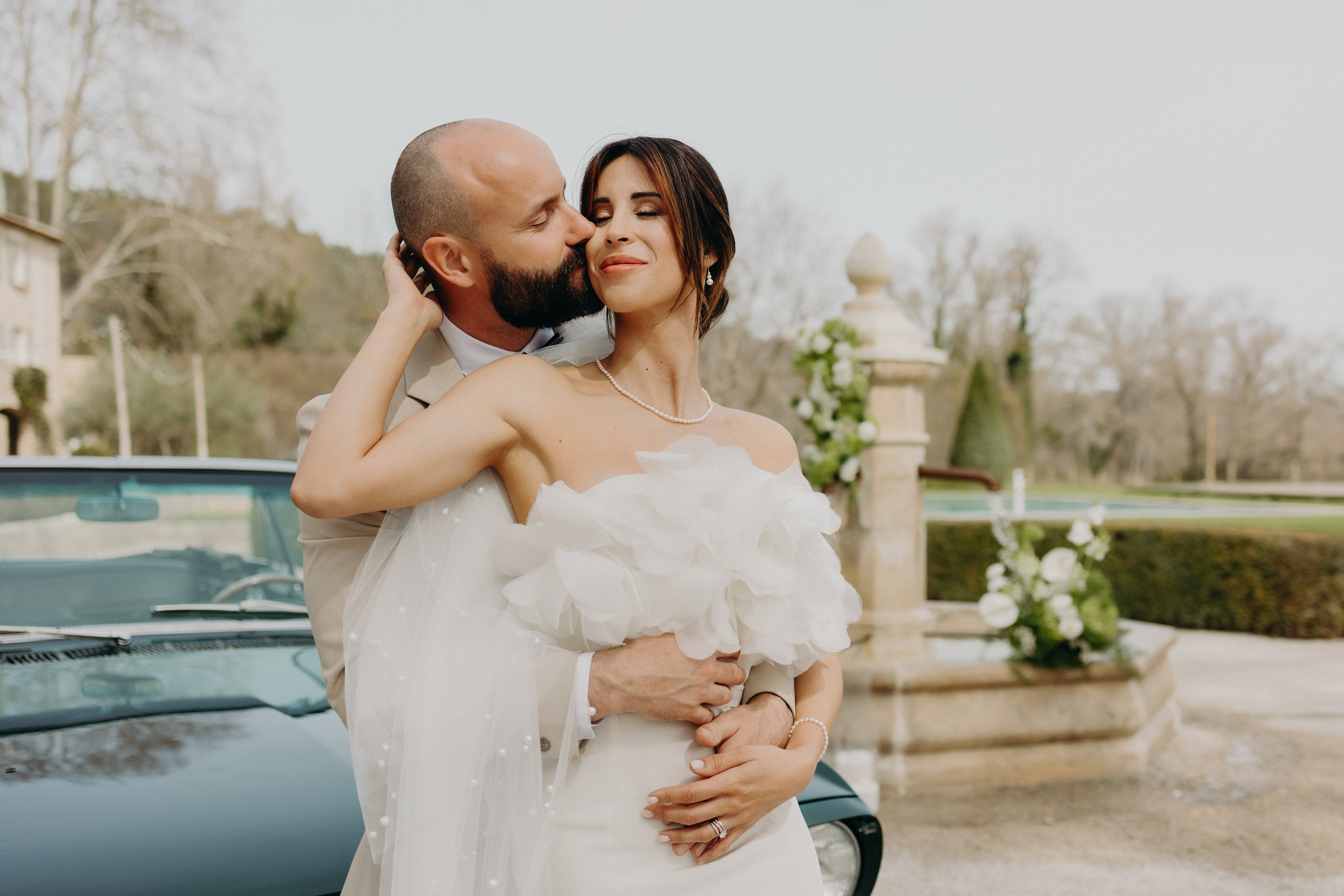 Un couple de jeunes mariés s’enlace tendrement devant une voiture vintage, sous une lumière douce, lors de leur réception de mariage au décor aux nuances matcha et olive, mêlant élégance naturelle et romantisme délicat.