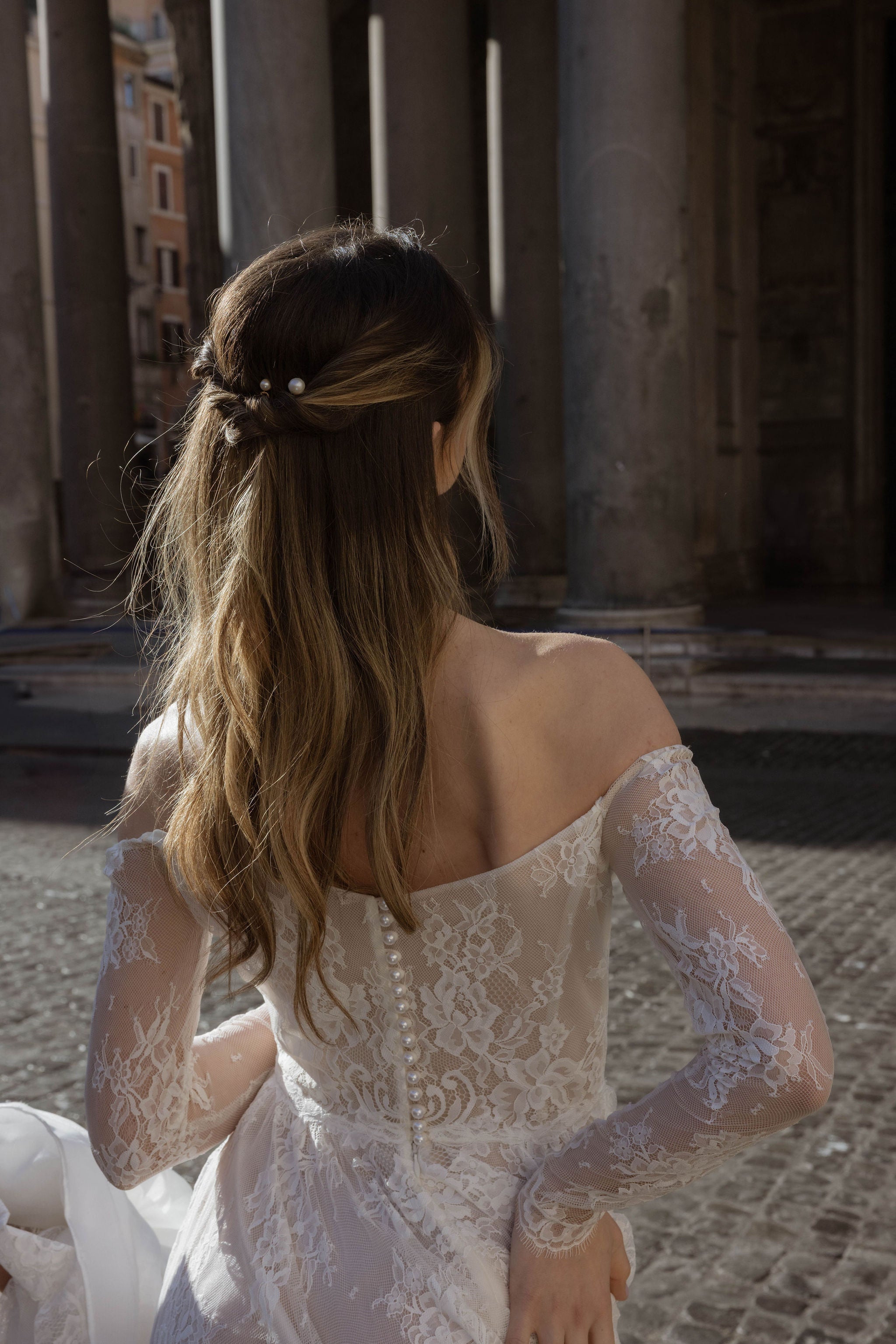 Mariée de dos portant une belle robe bustier en dentelle aux manches longues, avec une fermeture ornée de perles. Ses cheveux sont mi-lâchés et mi-attachés, décorés de pics en perles.