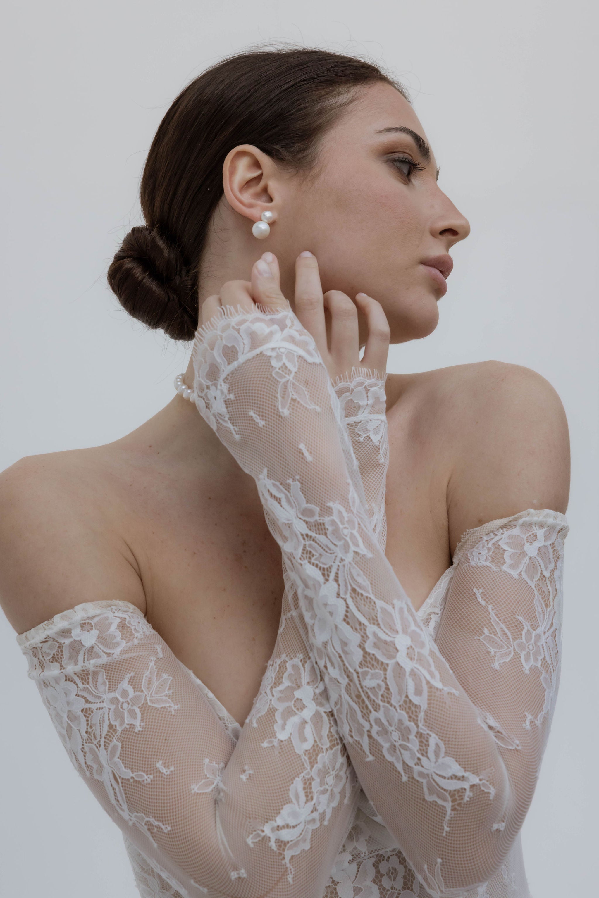 Mariée croisant les bras, portant une robe bustier avec manches en dentelle, un chignon parfaitement attaché, et des boucles d’oreilles à deux perles de tailles différentes.
