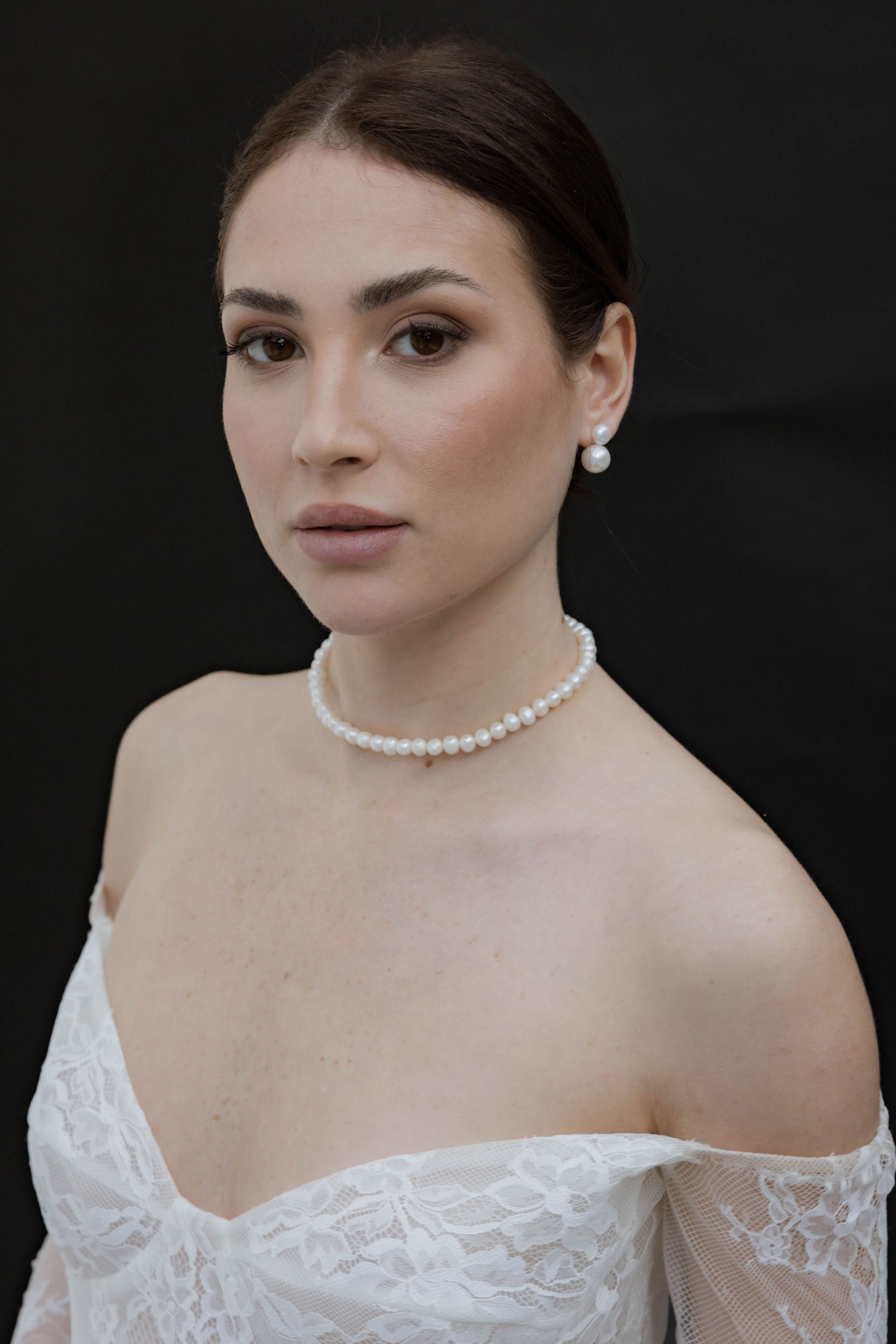 Mariée portant une jolie robe bustier en dentelle, un collier ras de cou en perles, ainsi que des boucles d’oreilles élégantes composées de deux perles.