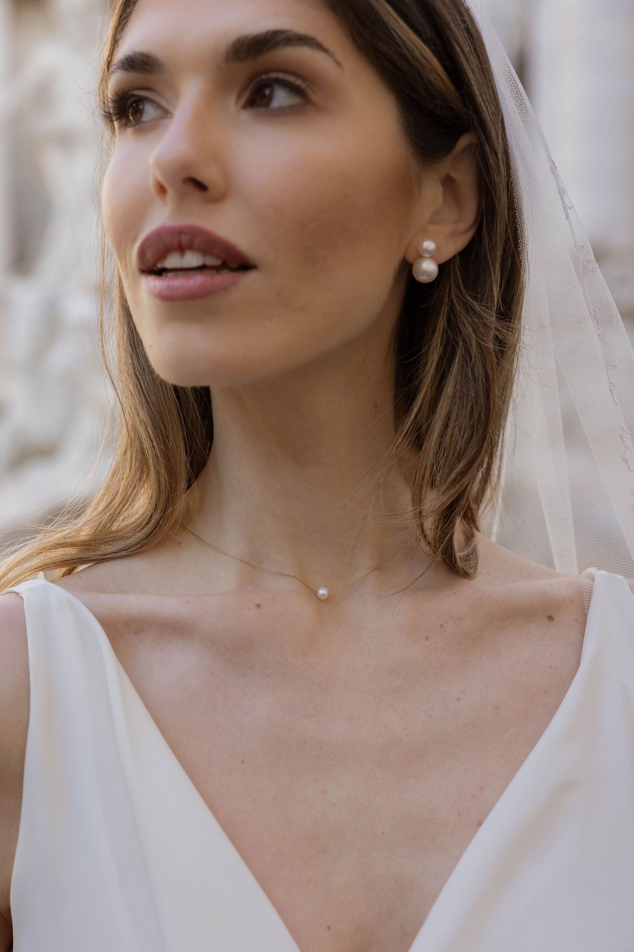 Mariée vue de près portant des boucles d’oreilles à deux perles de tailles différentes, un collier à chaîne fine avec une perle centrale, cheveux lâchés, décolleté en V et voile de mariée.