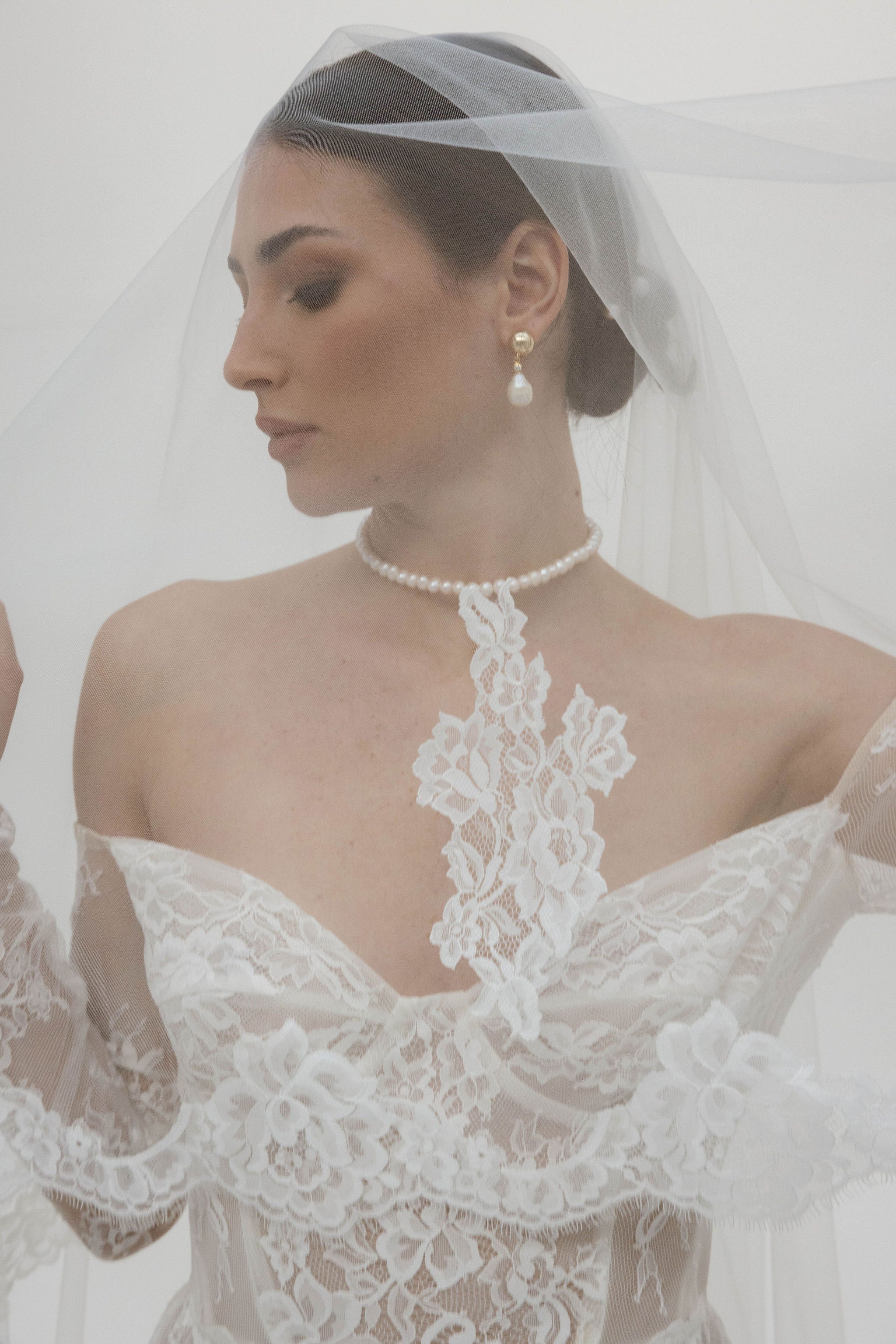 Mariée sous son voile en dentelle, coiffée d’un chignon élégant. Elle porte de jolies boucles d’oreilles clips ornées de perles, ainsi qu’un collier ras de cou en perles. Sa robe bustier en dentelle complète ce look raffiné et délicat.