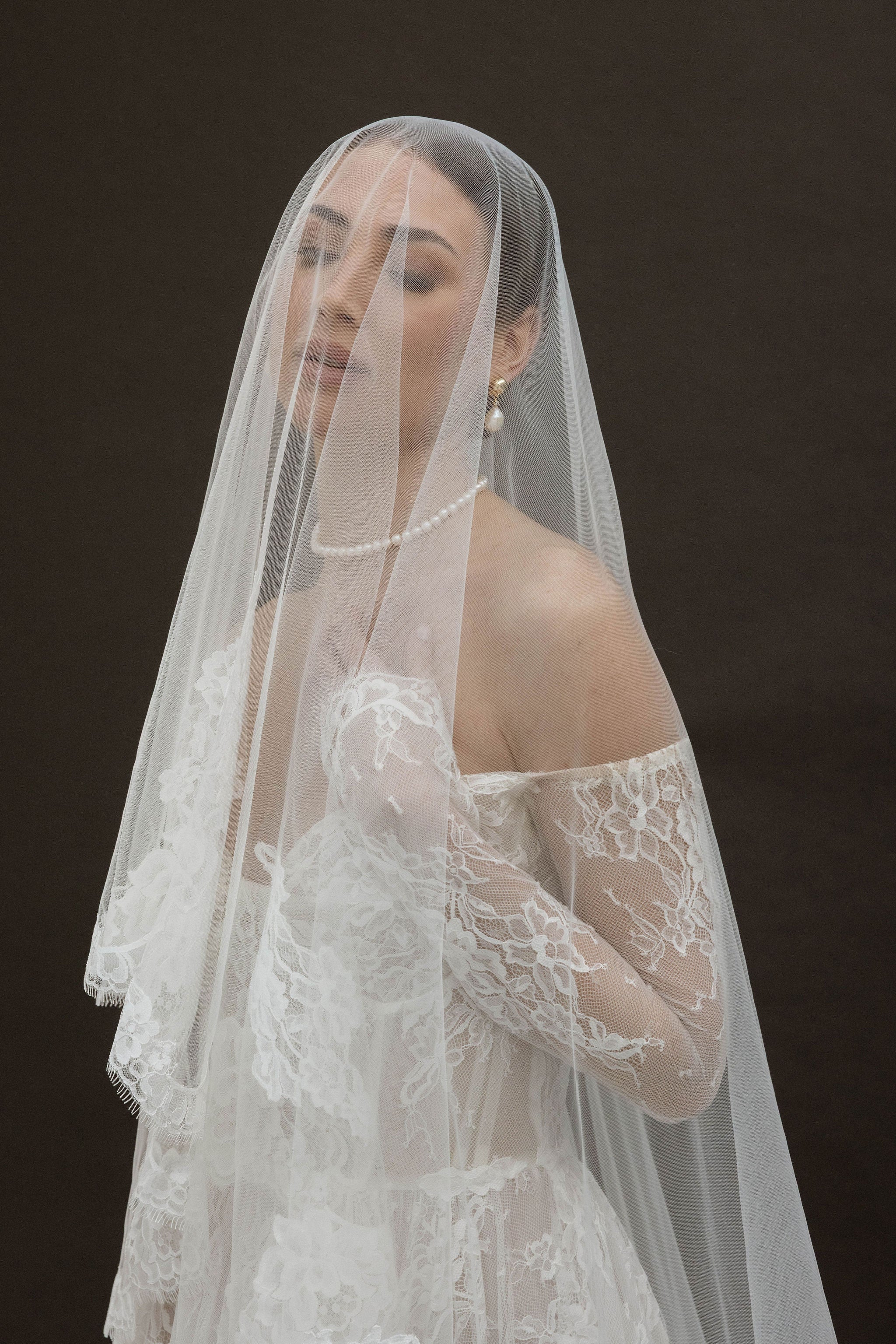 Mariée vue de dos, coiffée d’un chignon bas parfaitement réalisé, partiellement recouvert par un voile en dentelle fine. Elle porte un collier ras de cou en perles qui souligne délicatement la nuque, ainsi que des boucles d’oreilles clips élégantes, composées de deux perles de tailles différentes. Sa robe de mariée en dentelle dévoile de subtils détails dans le dos, pour un style à la fois romantique et raffiné