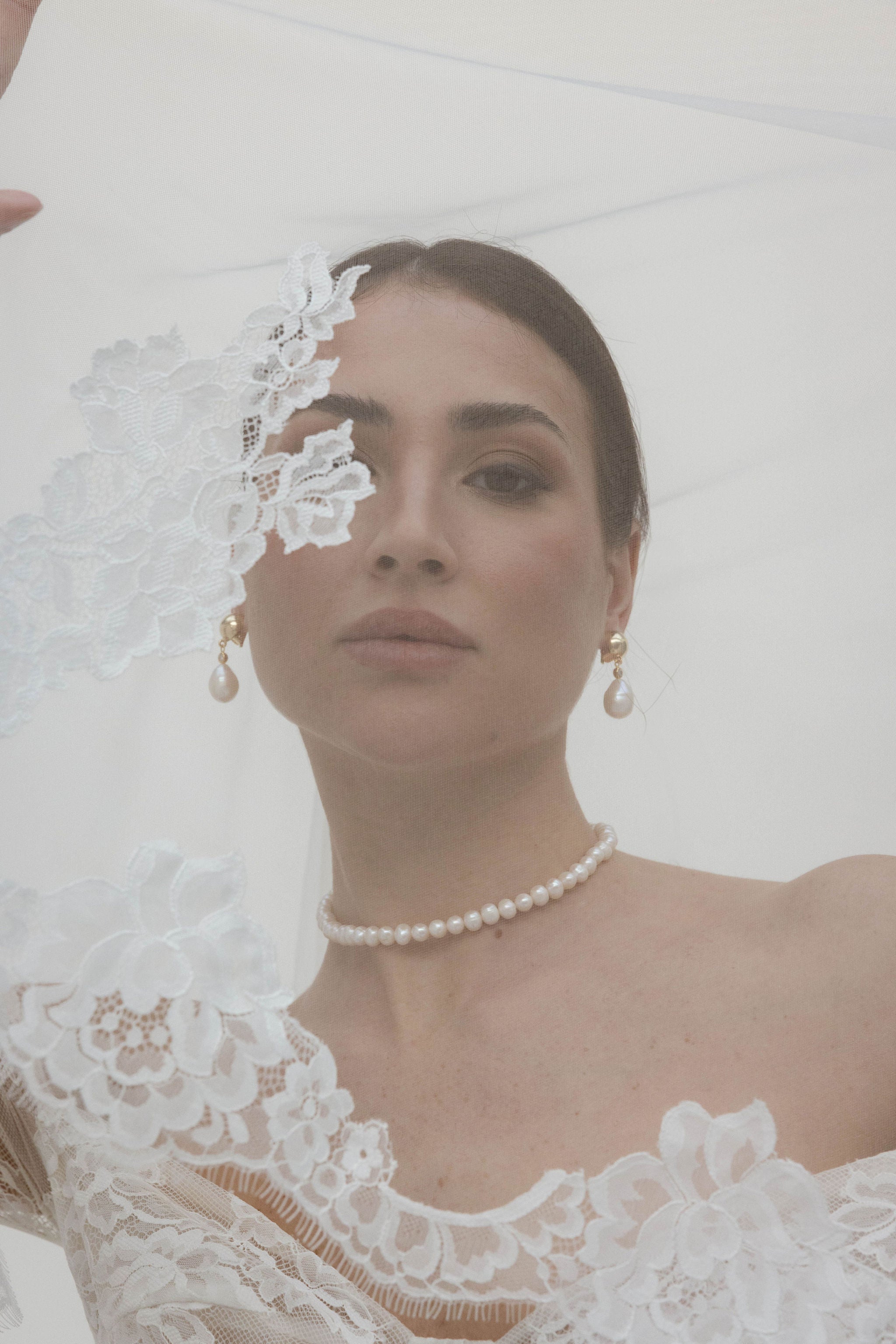 Mariée sous un voile en dentelle, portant un collier ras de cou en perles, des boucles d’oreilles en perles montées sur clips, une robe en dentelle, et les cheveux attachés en arrière dans une coiffure élégante.