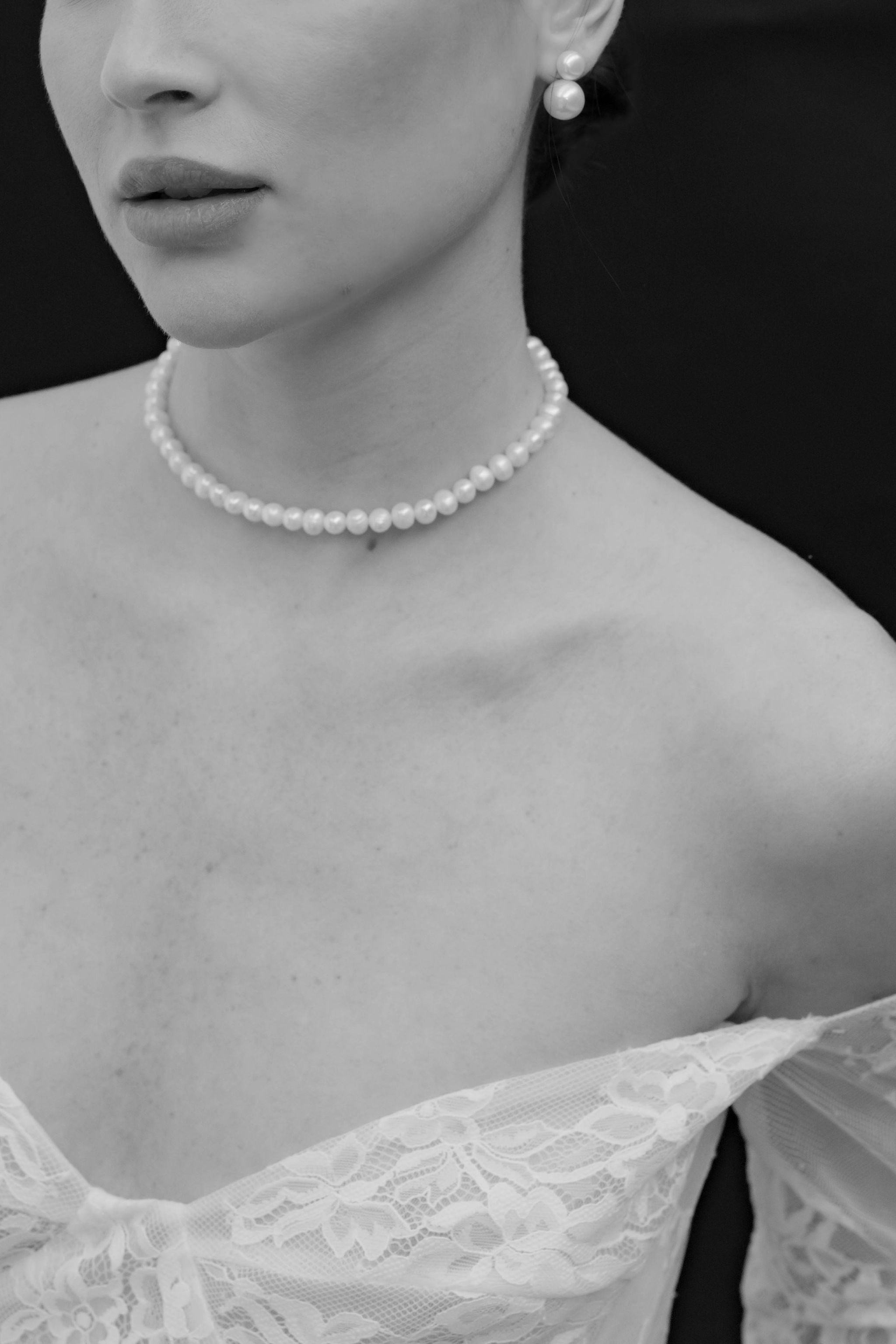 Vue rapprochée du cou d'une mariée sous un voile en dentelle, portant un collier ras de cou en perles, des boucles d’oreilles à clips ornées de perles, et un aperçu de sa robe en dentelle.
