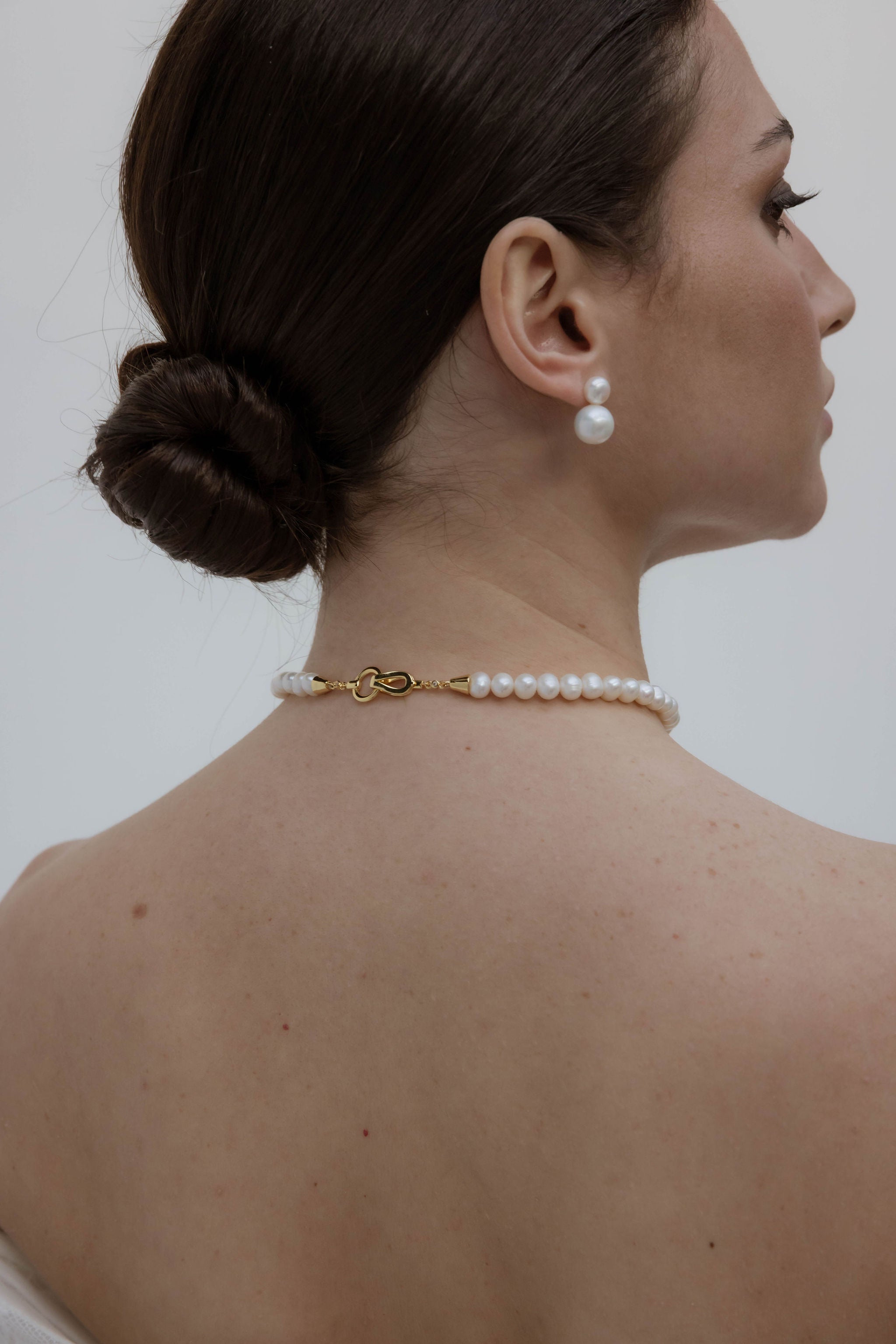 Vue de dos d’une mariée élégante avec un chignon bas soigneusement attaché, orné de boucles d’oreilles clips composées de deux perles, l’une petite et l’autre plus grosse.