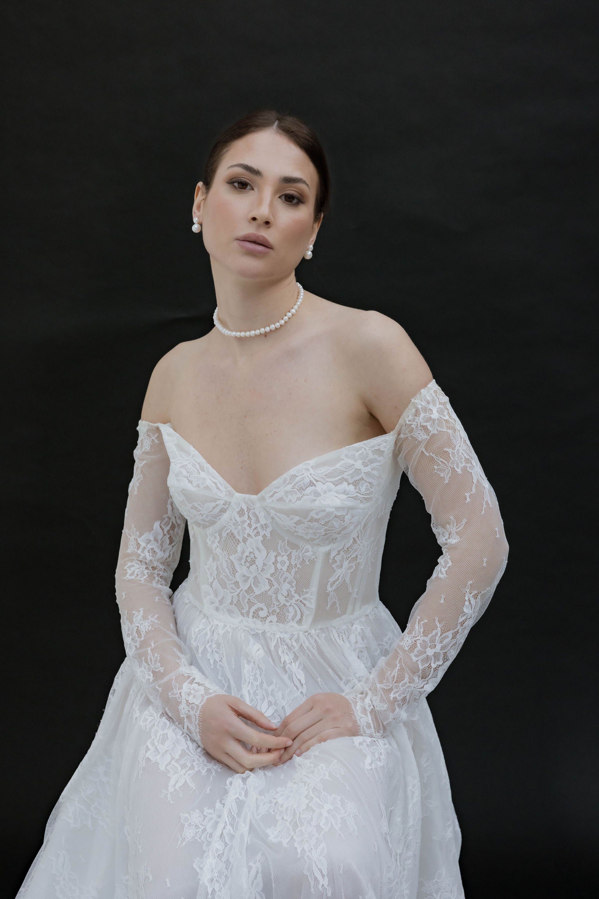 Mariée vue de dos, les cheveux relevés en un chignon bas élégant mettant en valeur un collier ras de cou en perles délicates. Elle porte de jolies boucles d’oreilles clips, composées de deux perles de tailles différentes. Sa robe en dentelle dévoile subtilement le haut du dos, ajoutant une touche romantique et intemporelle à son allure.