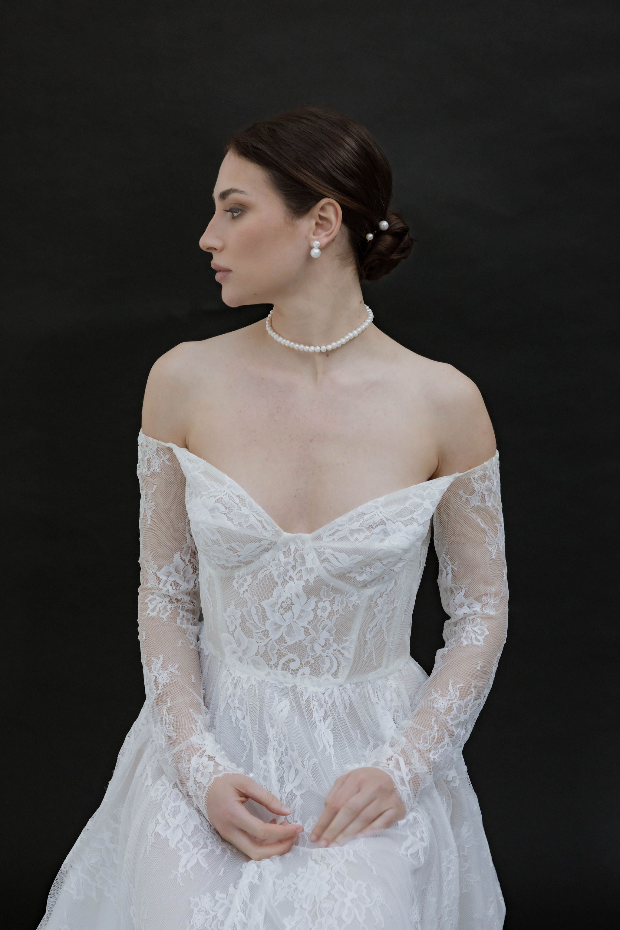 Mariée de dos dans une lumière douce, les cheveux soigneusement attachés en un chignon flou. Autour de son cou, un collier ras de cou en perles apporte une touche classique, tandis que ses boucles d’oreilles clips avec deux perles, l’une discrète et l’autre plus généreuse, captent la lumière avec élégance. Sa robe en dentelle aux détails fins complète cette silhouette à la fois moderne et romantique.