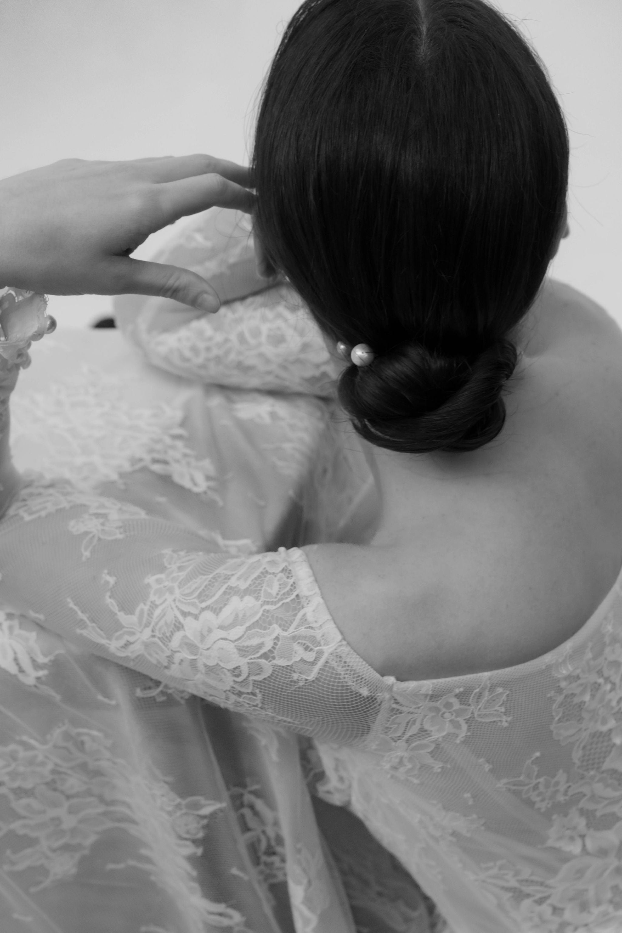 Mariée vue de haut, portant un chignon bien tiré orné d’un pic de perles sur le côté, habillée d’une robe en dentelle à manches longues.