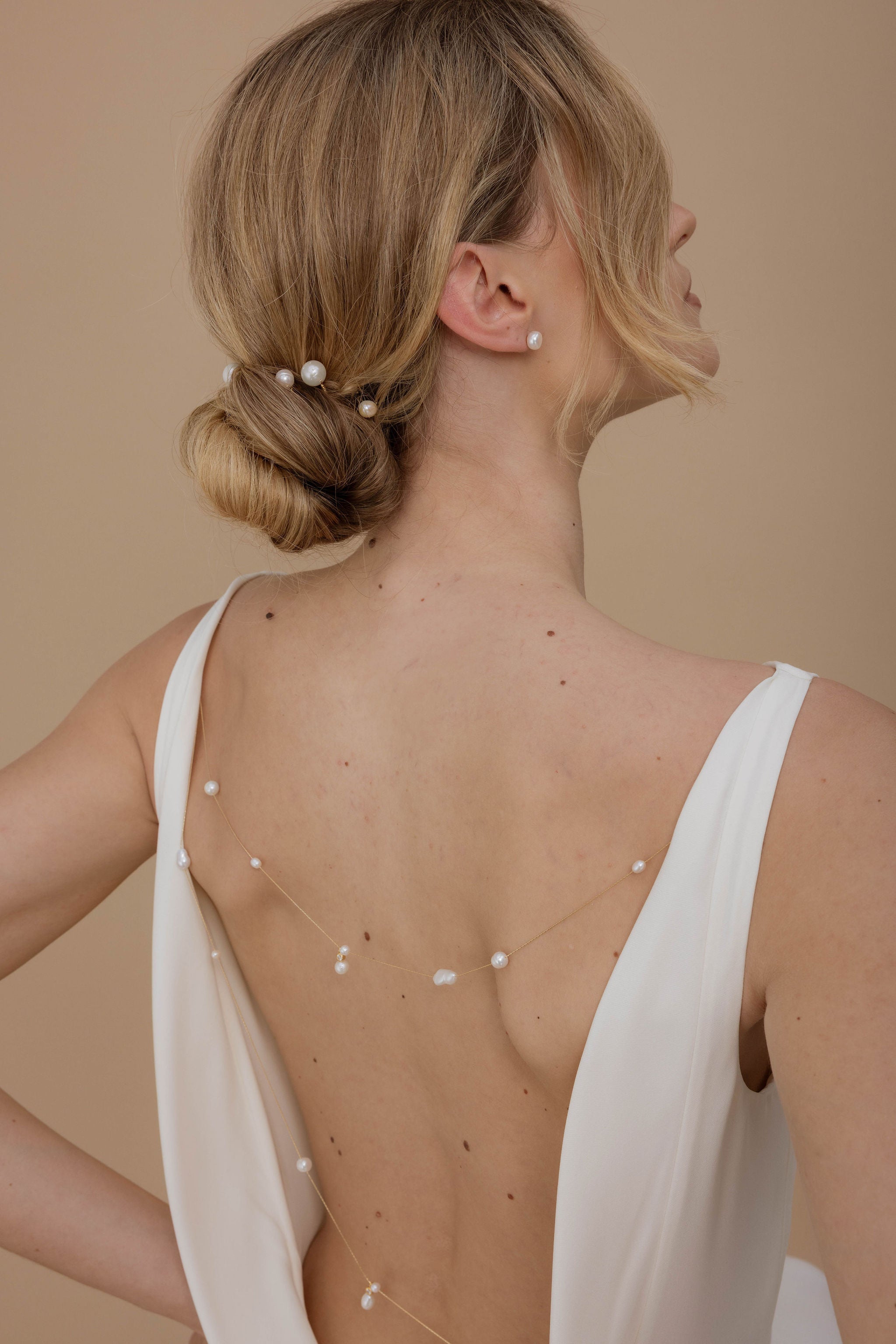 Mariée élégante avec un joli chignon orné de pics en perles naturelles, portant un décolleté de dos souligné par un collier de robe délicat avec de belles perles, accompagnée de boucles d’oreilles en perles assorties.