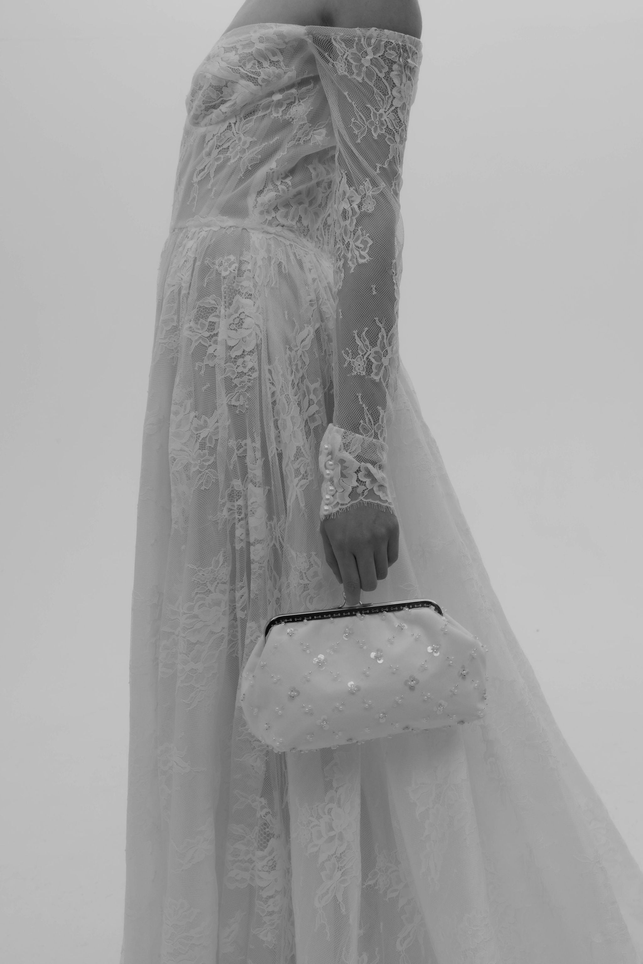 Mariée portant une jolie robe bustier en dentelle avec manches longues ornées de perles, tenant un sac de mariée décoré de brillants au bout des doigts.