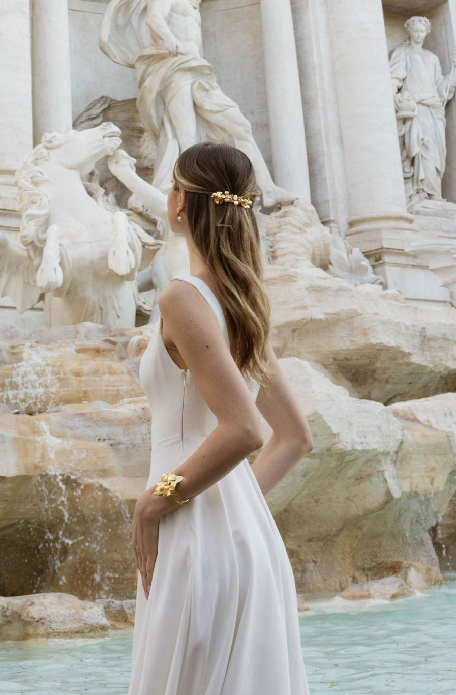 Mariée devant une fontaine, portant une jolie robe blanche et un beau peigne floral dans ses cheveux, regard tourné vers la fontaine