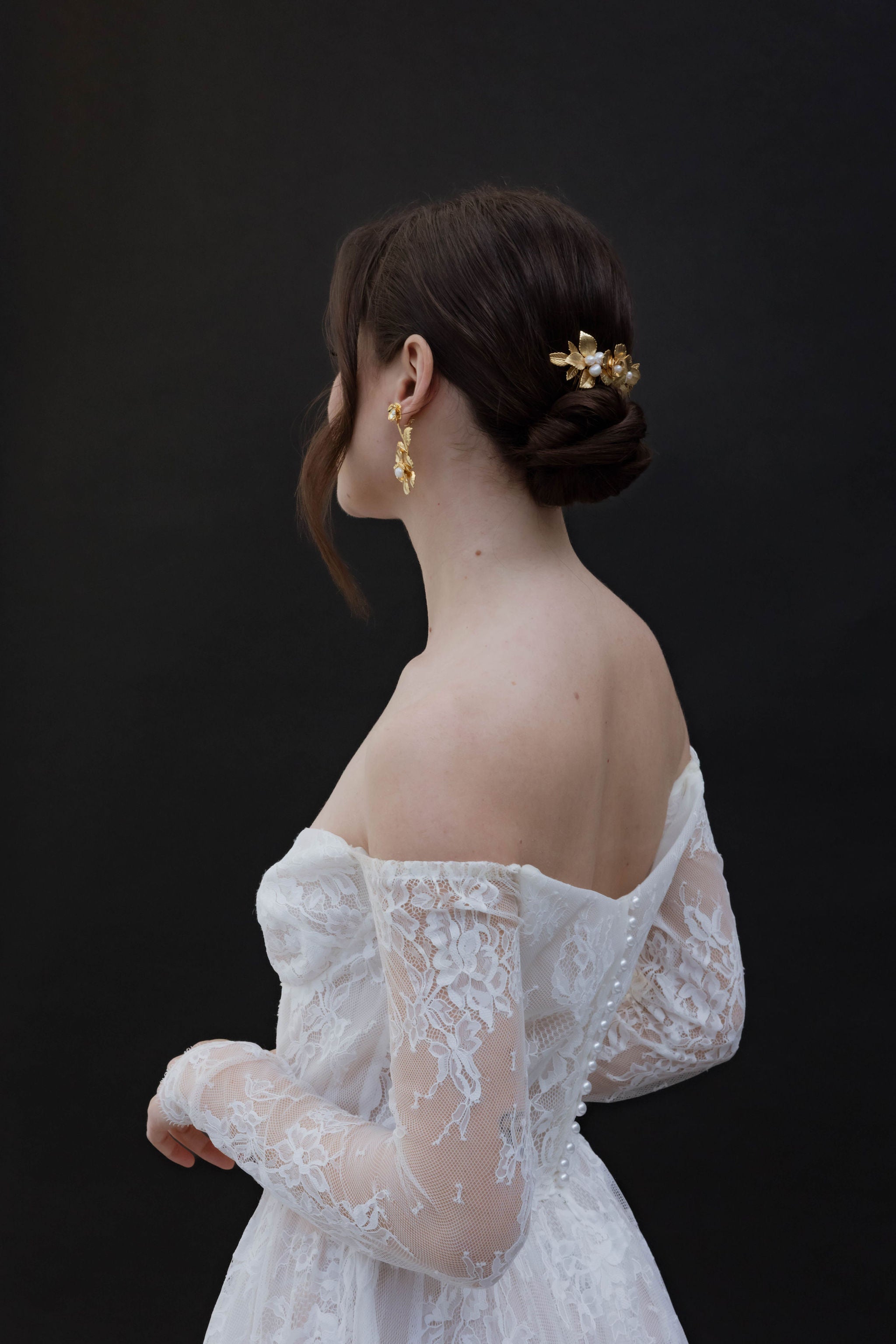 Mariée avec un peigne floral orné de perles, chignon bas et une mèche délicate tombant devant, portant une jolie robe en dentelle et des boucles d'oreilles assorties pour un look élégant et harmonieux.