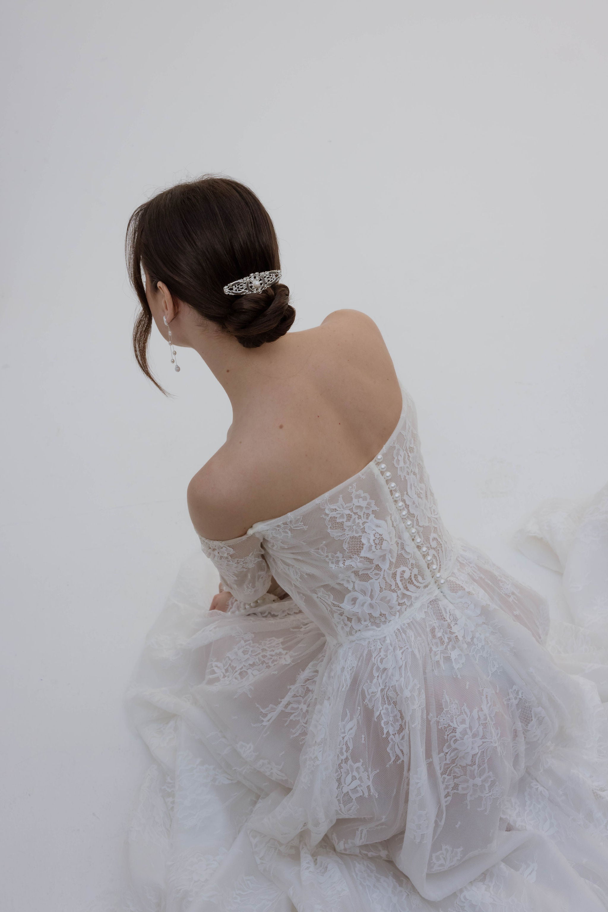 Mariée vue de dos, coiffée d’un chignon bas orné d’un pic romantique, portant une robe de mariage en dentelle avec manches longues et bustier.