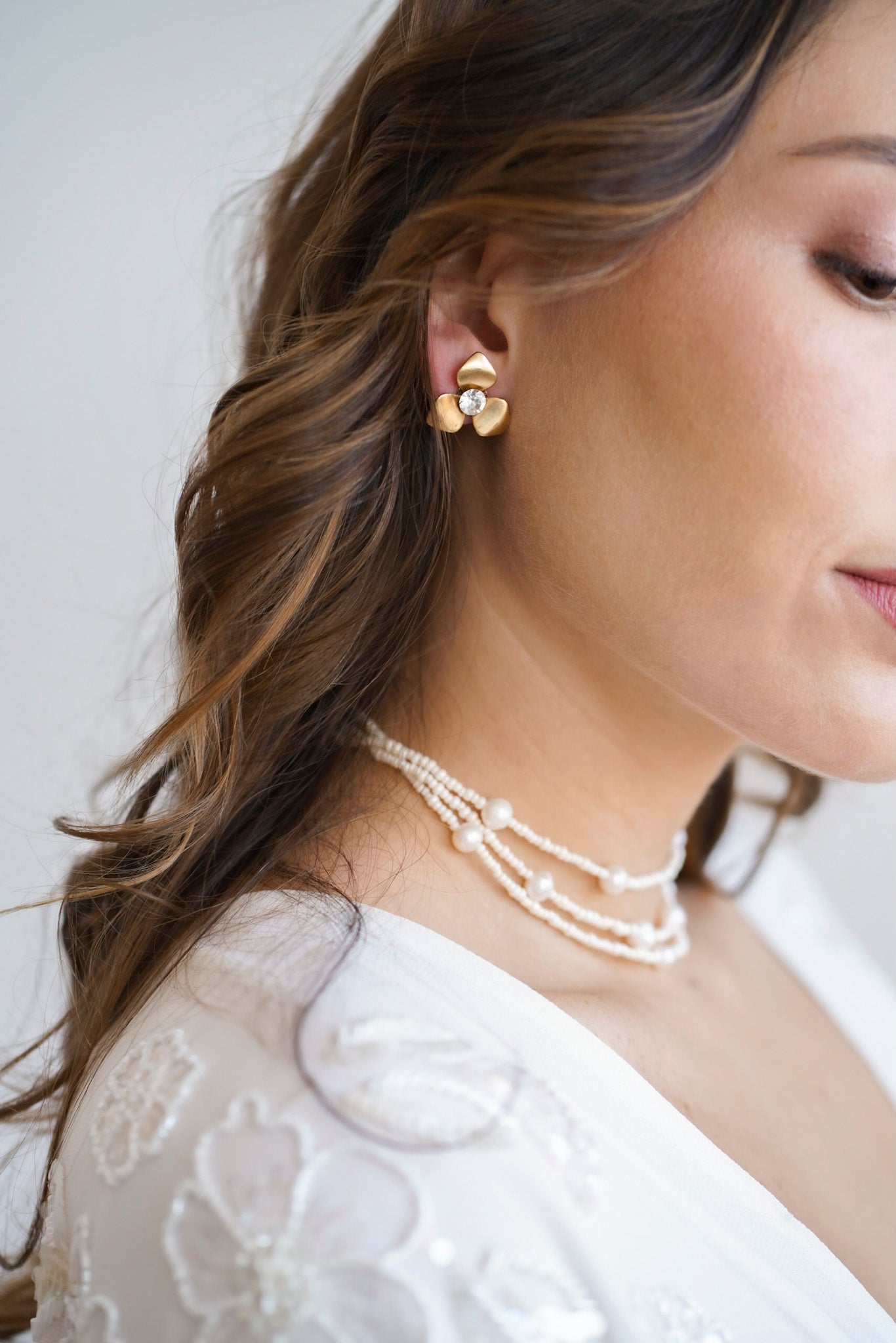 Mariée avec de long cheveux ondulés au style bohème portant des boucles d'oreilles dorées en forme de fleur avec un cristaux Swarovski en sont centre et un collier ras-de-cou à trois rangées en perles blanches