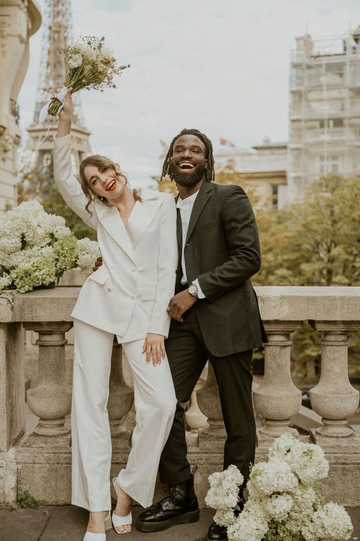 Couple derrière la tour Eiffel après leur mariage civl, ils sourient et la mariée porte un pantalon et un blazer de mariage