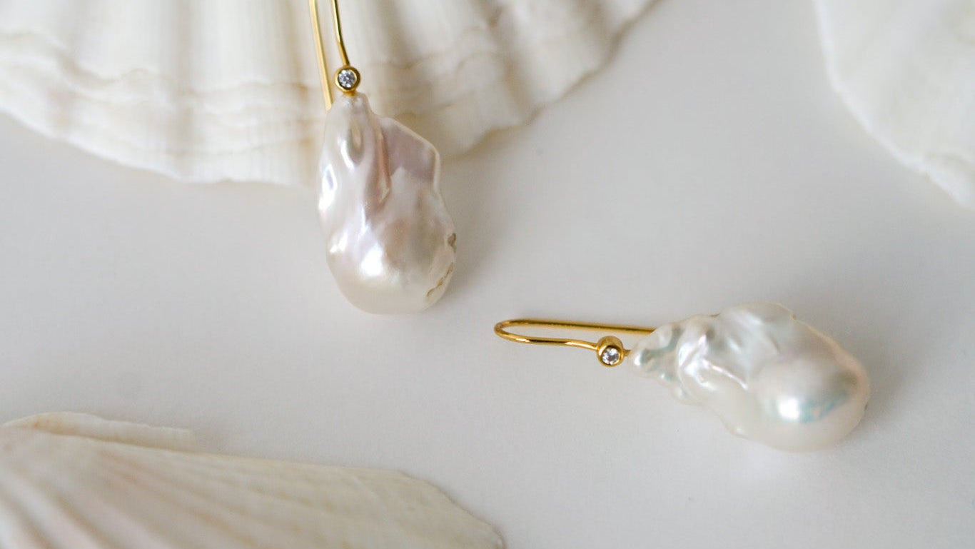 boucles d'oreilles avec une perle naturelle baroque irrégulière pour mariage et son attache dorée sur une table blanche avec des coquillages en fond