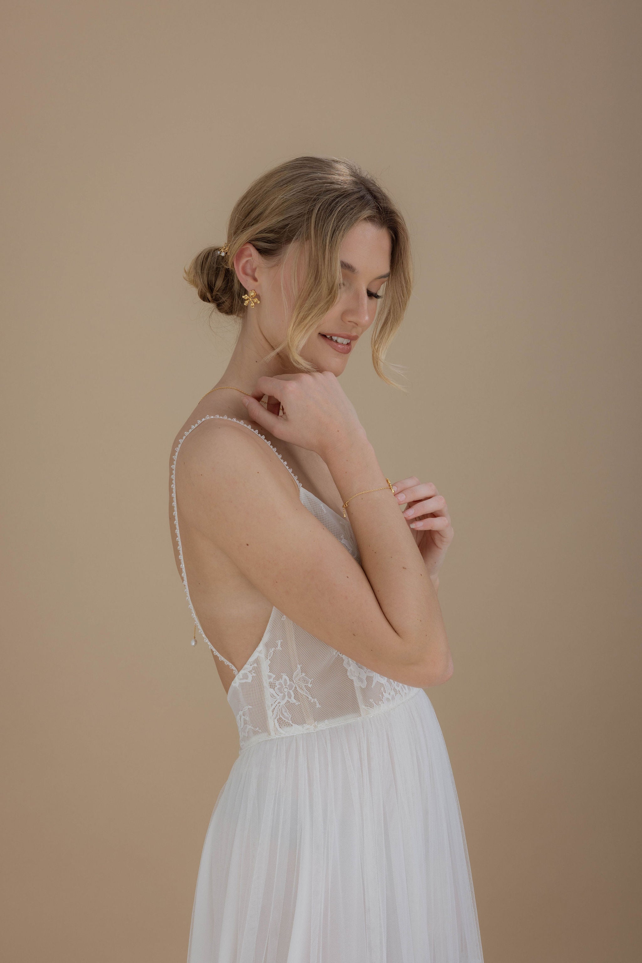 Mariée en robe blanche élégante à décolleté dos nu, coiffée d’un chignon sophistiqué, portant des boucles d'oreilles courtes florales en or, pour un look romantique et raffiné