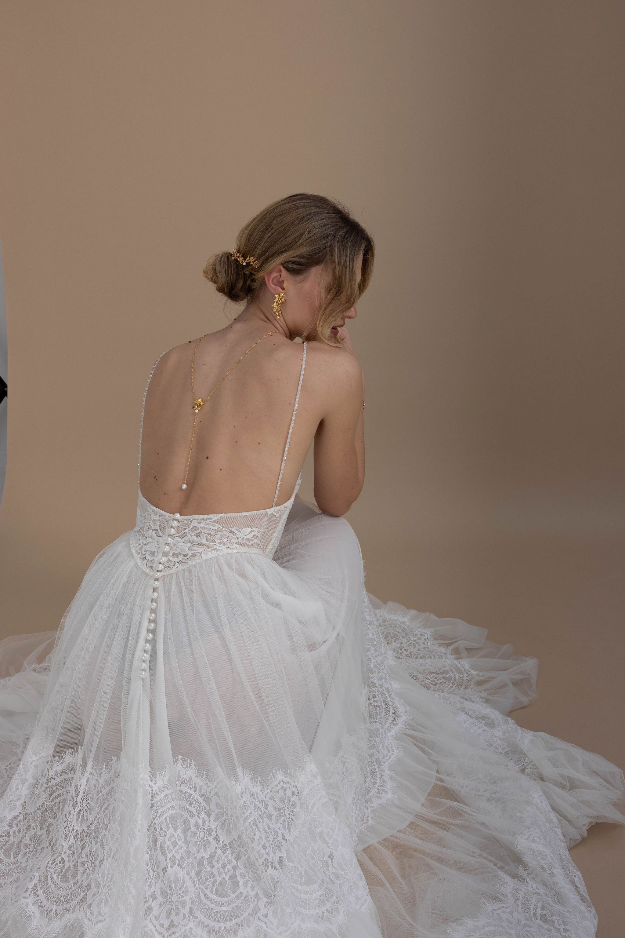 Mariée en robe en dentelle dos nu, coiffée d’un chignon bas élégant, portant un collier de dos floral avec des perles naturelles et des accessoires cheveux délicats pour une allure romantique.