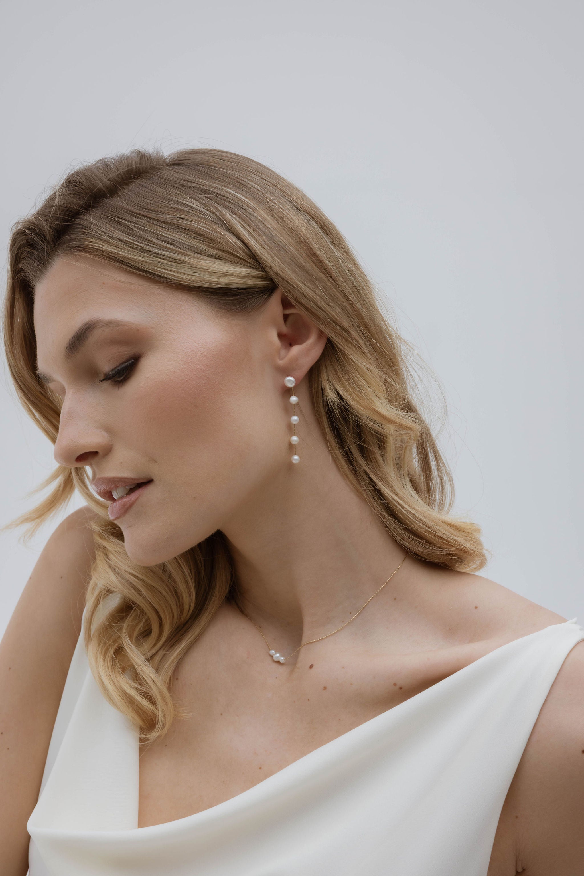Une mariée élégante dans sa robe blanche, aux cheveux lâchés et parée de bijoux en perles naturelles, créant un look à la fois simple et sophistiqué