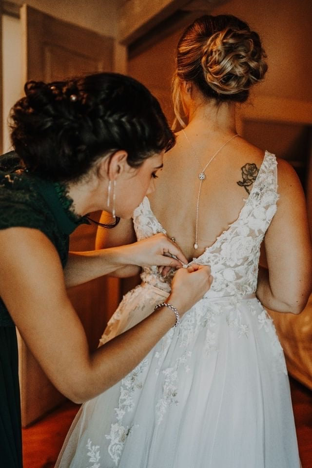 Jeune Mariée qui met sa robe de mariée avec l'aide de sa demoiselle d'honneur montrant son collier de dos en zircons
