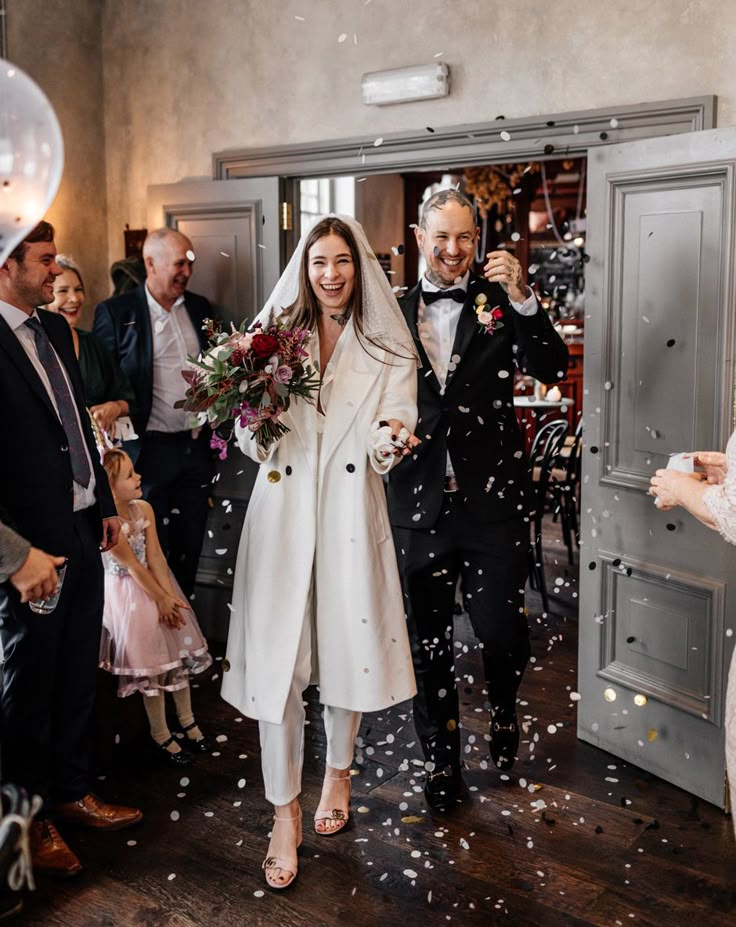 Couple de mariées qui sortent de la mairie, la mariée porte un tailleur blanc et un voile ainsi qu'un bouquet de mariée