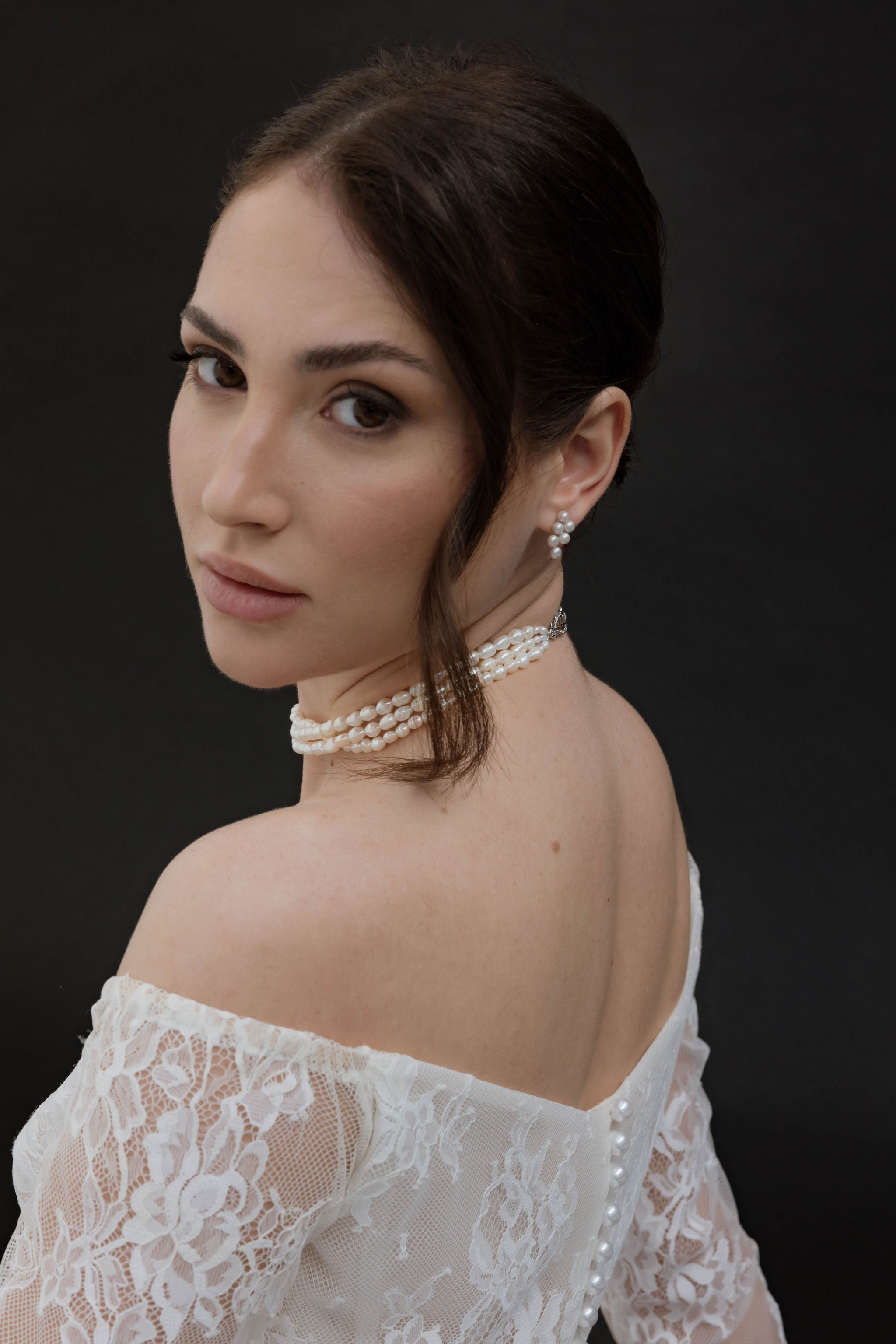 Mariée de face avec un chignon bas, portant des boucles d’oreilles à six perles et un collier en perles à trois rangs, style minimaliste et raffiné.