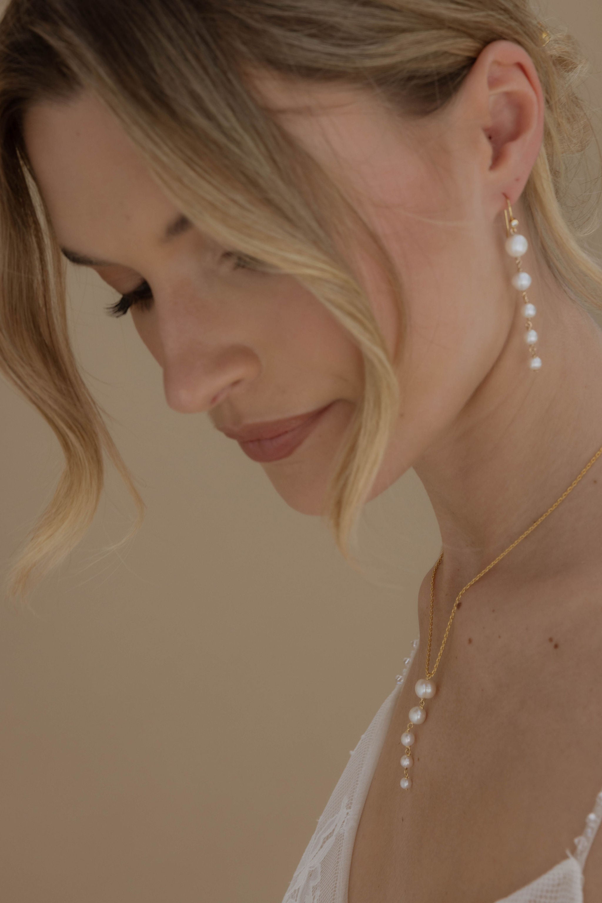 Mariée en robe à bretelles, coiffée d’un chignon, portant des boucles d’oreilles pendantes avec cinq perles de la plus grande à la plus petite et un collier assorti.