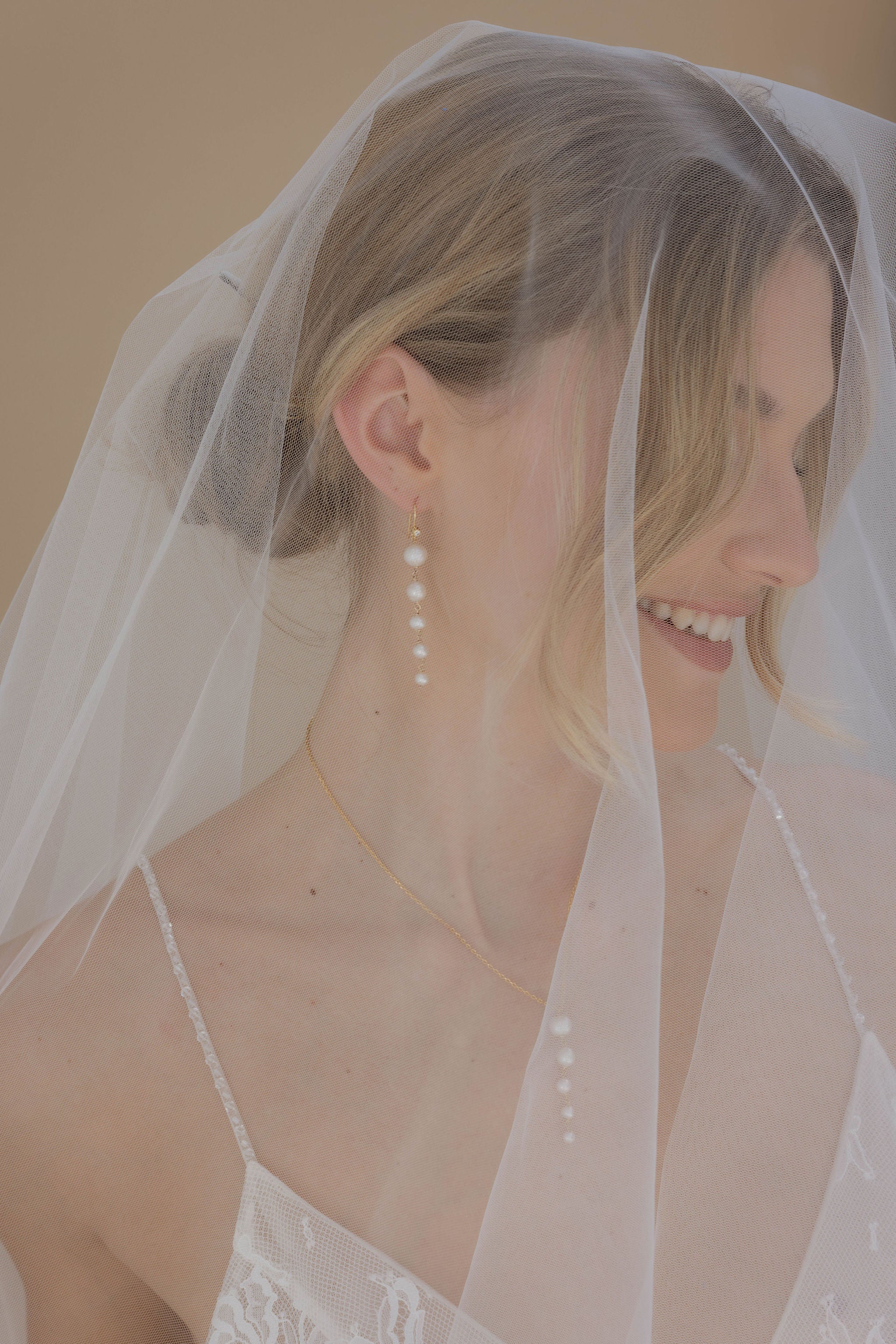 Mariée avec un voile, une robe à bretelles et un chignon. Elle porte des boucles d’oreilles avec cinq perles de tailles différentes et un collier assorti.
