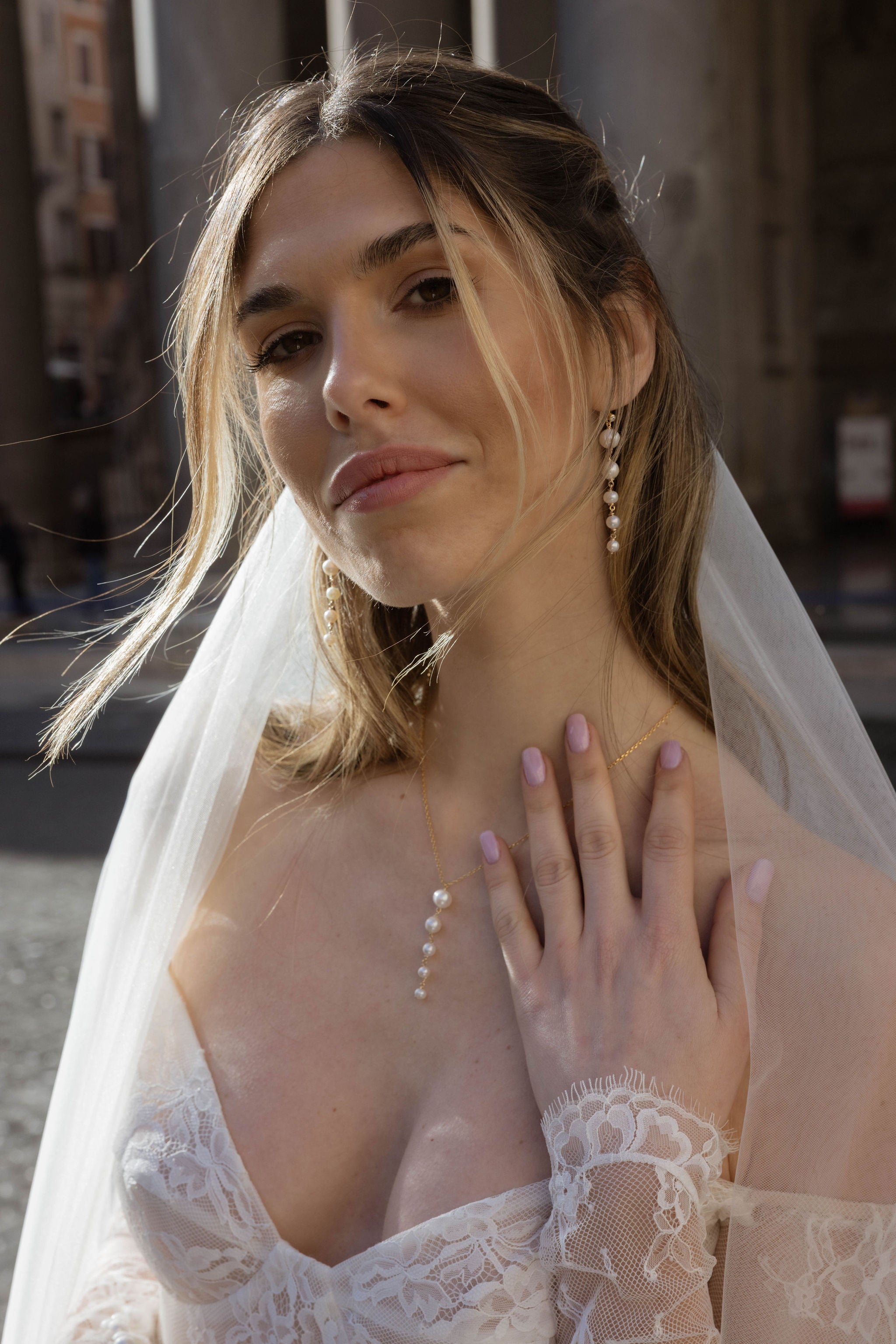 Mariée en extérieur, portant une belle robe en dentelle, des boucles d’oreilles assorties, un voile blanc et les cheveux détachés.