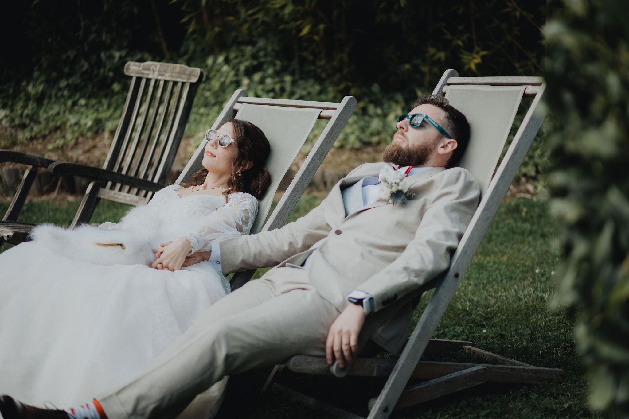 couple de jeunes mariés se tenant la main sur un transat avec des lunettes de soleil le jour de leur mariage