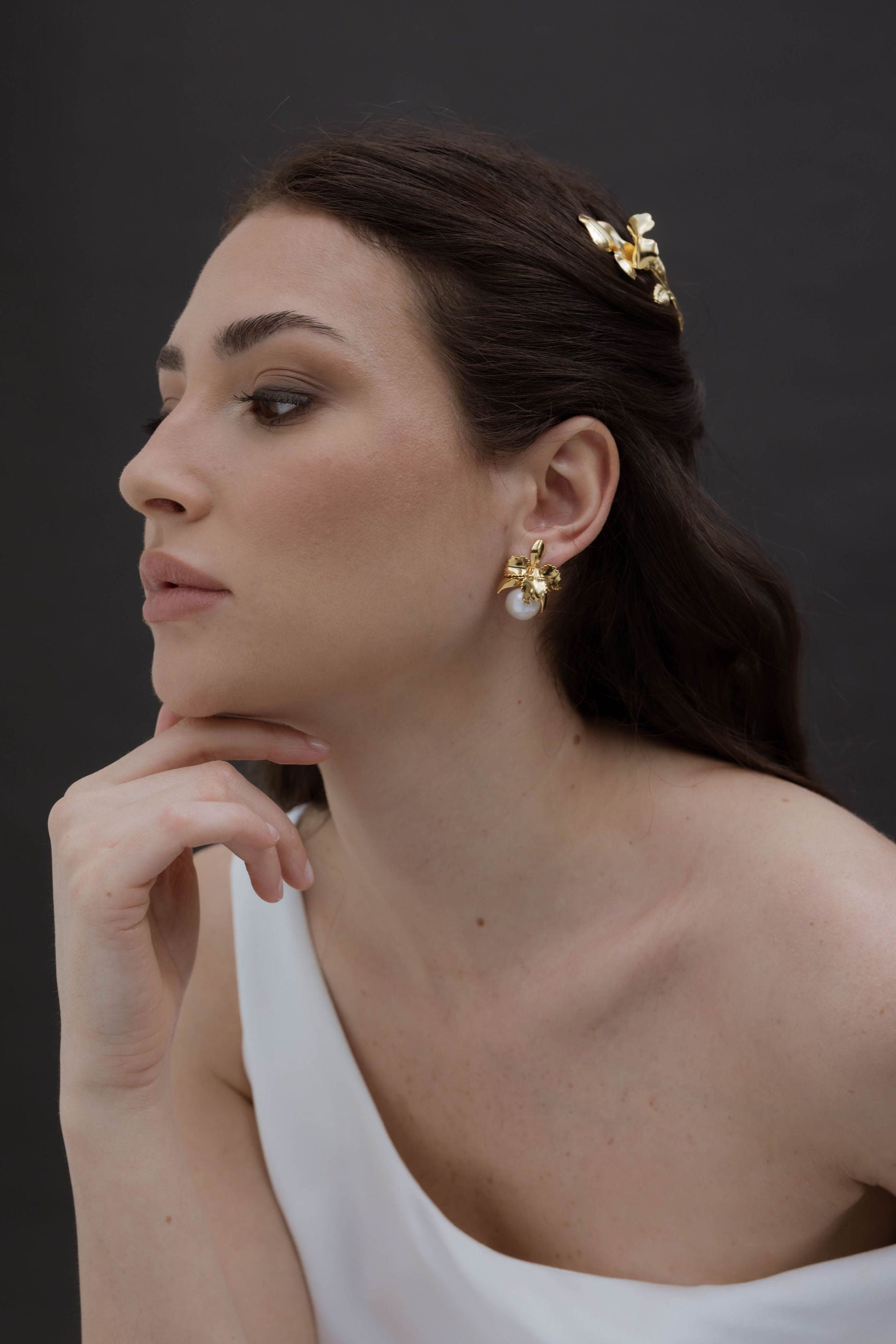 Mariée élégante en robe blanche asymétrique, les cheveux lâchés délicatement coiffés avec un accessoire raffiné. Elle porte des boucles d’oreilles florales délicates agrémentées d’une perle, ajoutant une touche de douceur et de féminité à son look.