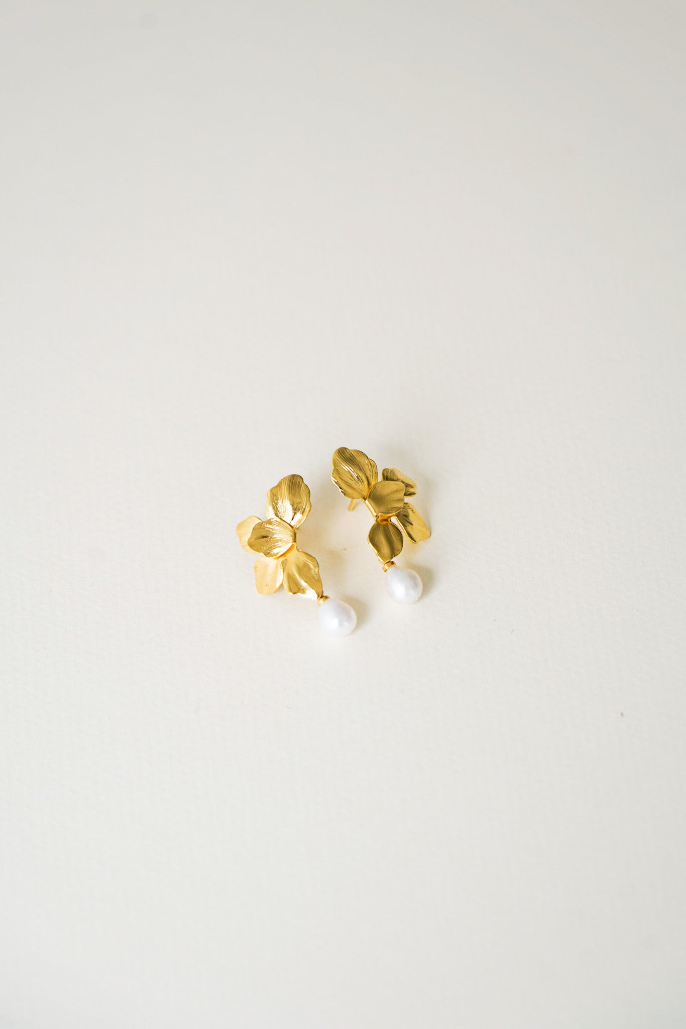 Paire de boucles d’oreilles dorées en forme de fleur délicatement sculptée, chaque pétale finement découpé laissant apparaître une perle naturelle en suspension à l’extrémité. Un design élégant et raffiné, idéal pour une mariée en quête de douceur et de poésie.