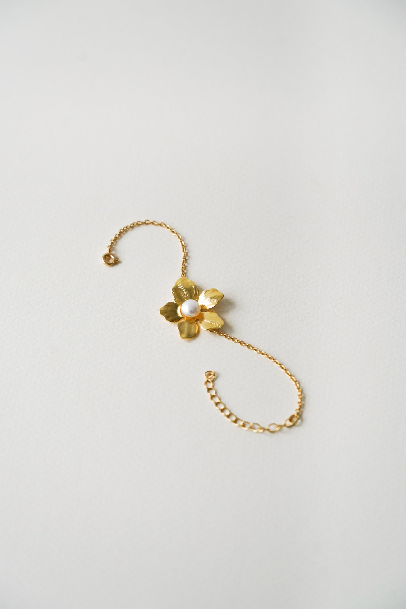 Bracelet raffiné en plaqué or, délicatement posé, mettant en valeur une élégante fleur à cinq pétales finement travaillés dans un ton doré lumineux. En son centre, une perle de culture blanche, ronde et éclatante, apporte une touche de douceur et de sophistication. Le bijou repose sur un fond neutre, soulignant son allure romantique et précieuse, idéale pour une mariée ou une occasion élégante.