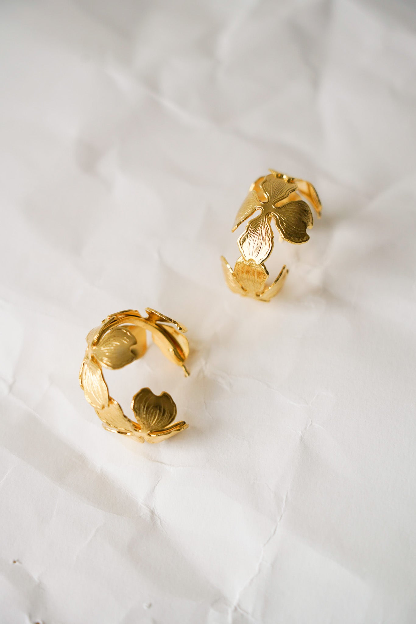 Créoles dorées délicates posées à plat, ornées de trois fleurs stylisées réparties le long du cercle. Chaque fleur est composée de quatre pétales légèrement arrondis avec de fins traits ciselés qui ajoutent texture et relief. Un bijou élégant et raffiné, parfait pour une touche florale et poétique.