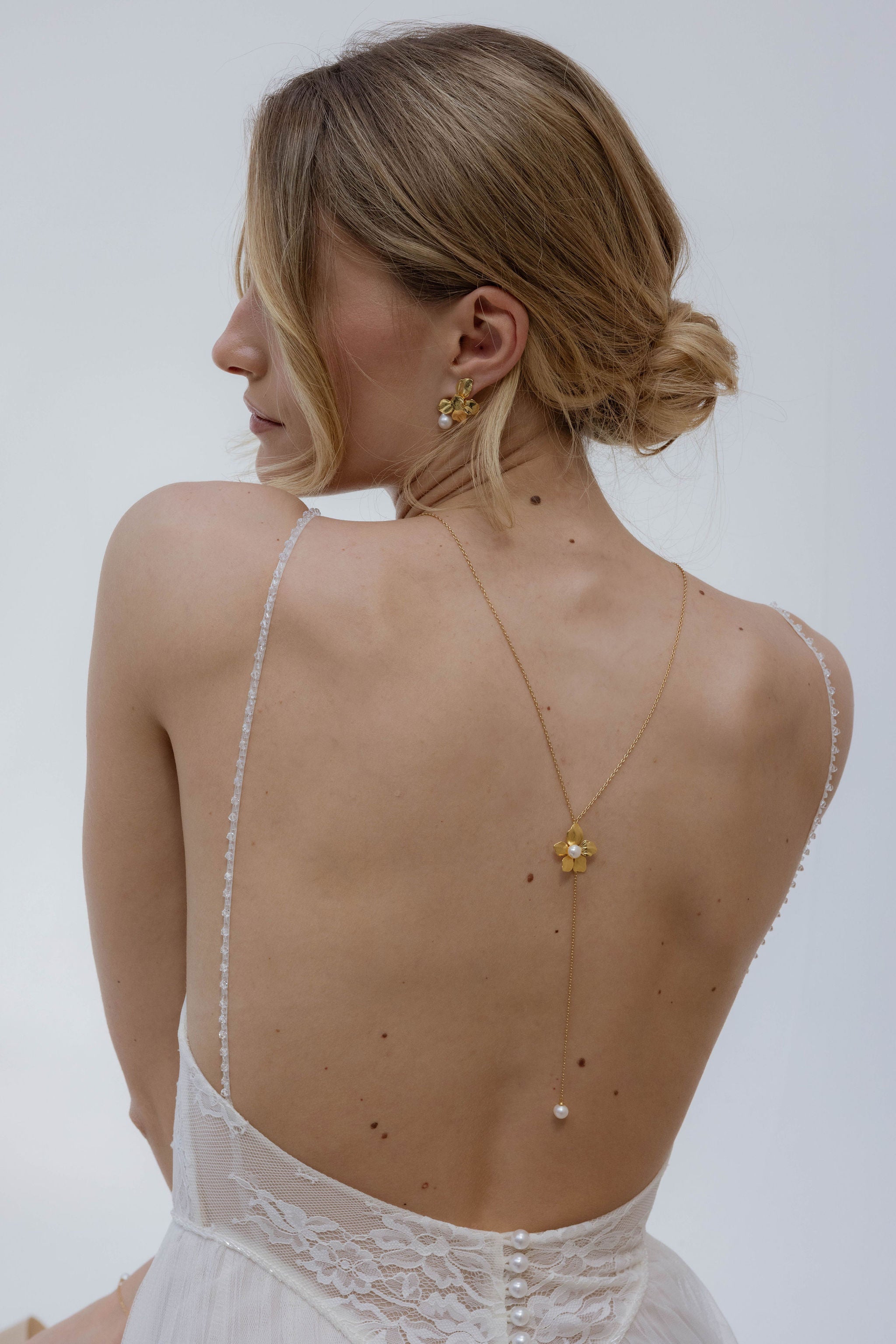 Mariée élégante de dos, avec une robe avec un jolie décolleté de dos et de la dentelle.
Portant un ensemble de bijoux en perles : boucles d’oreilles fines et bracelet assorti, parfaitement mis en valeur par un chignon bas travaillé.