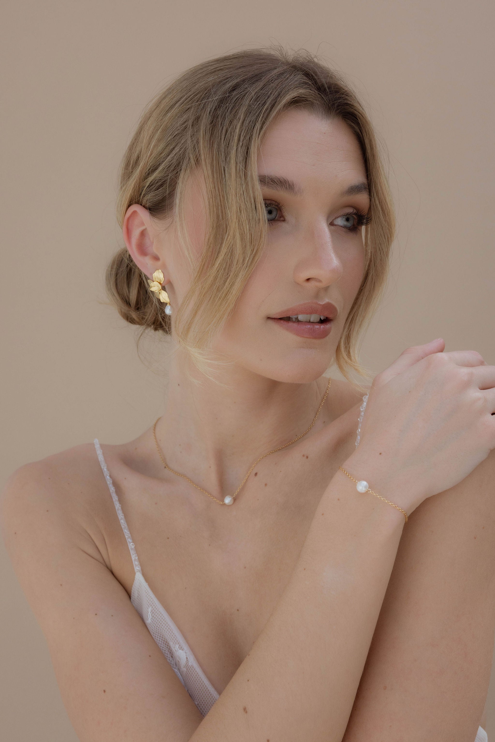 Mariée de profil coiffée d’un chignon bas, portant de délicates boucles d’oreilles pendantes avec perles blanches nacrées, assorties à un bracelet fin orné des mêmes perles.