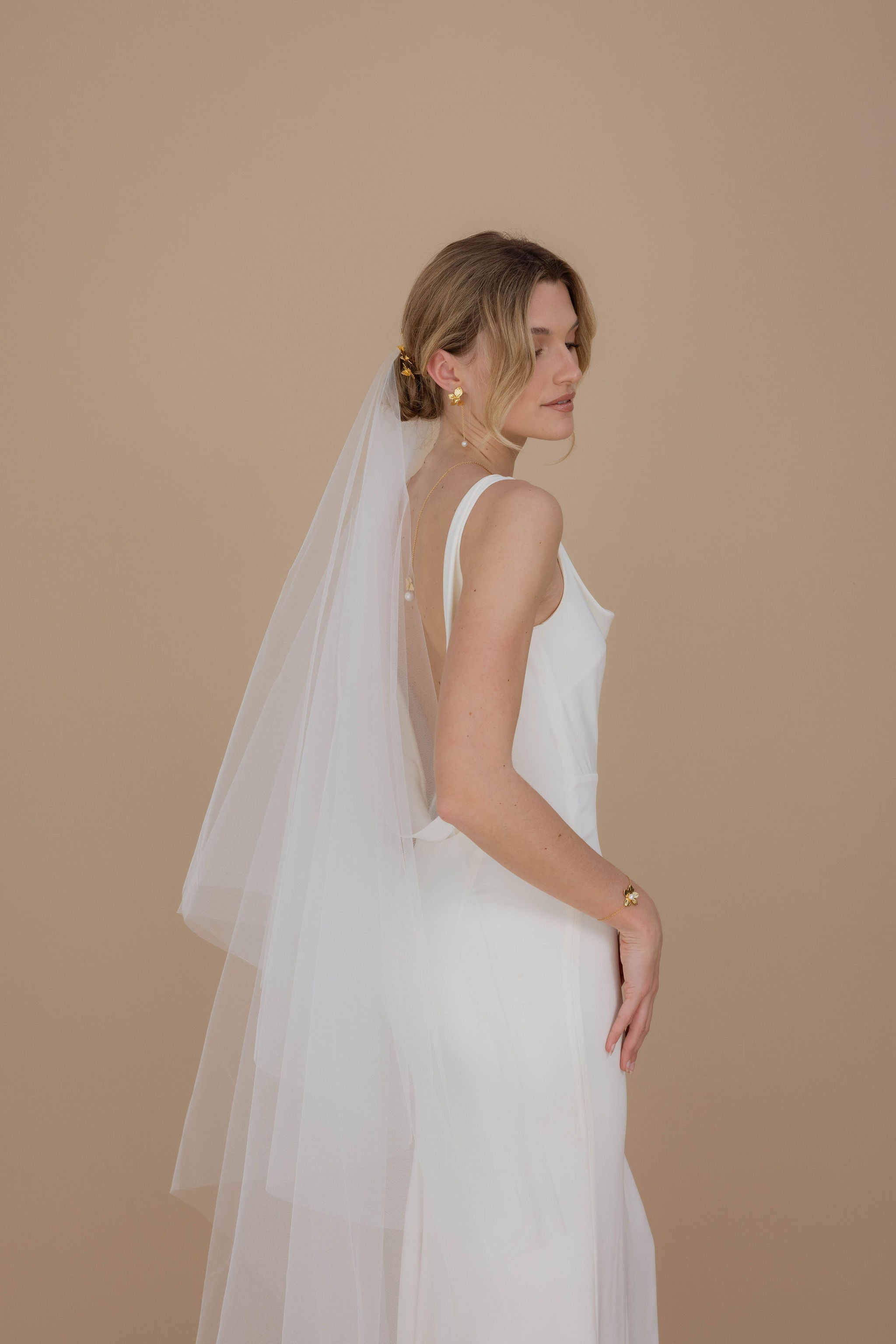 Vue arrière d’une mariée en robe blanche avec décolleté dos nu, accessoires de mariage avec fleurs et perles, chignon bas et voile vaporeux.