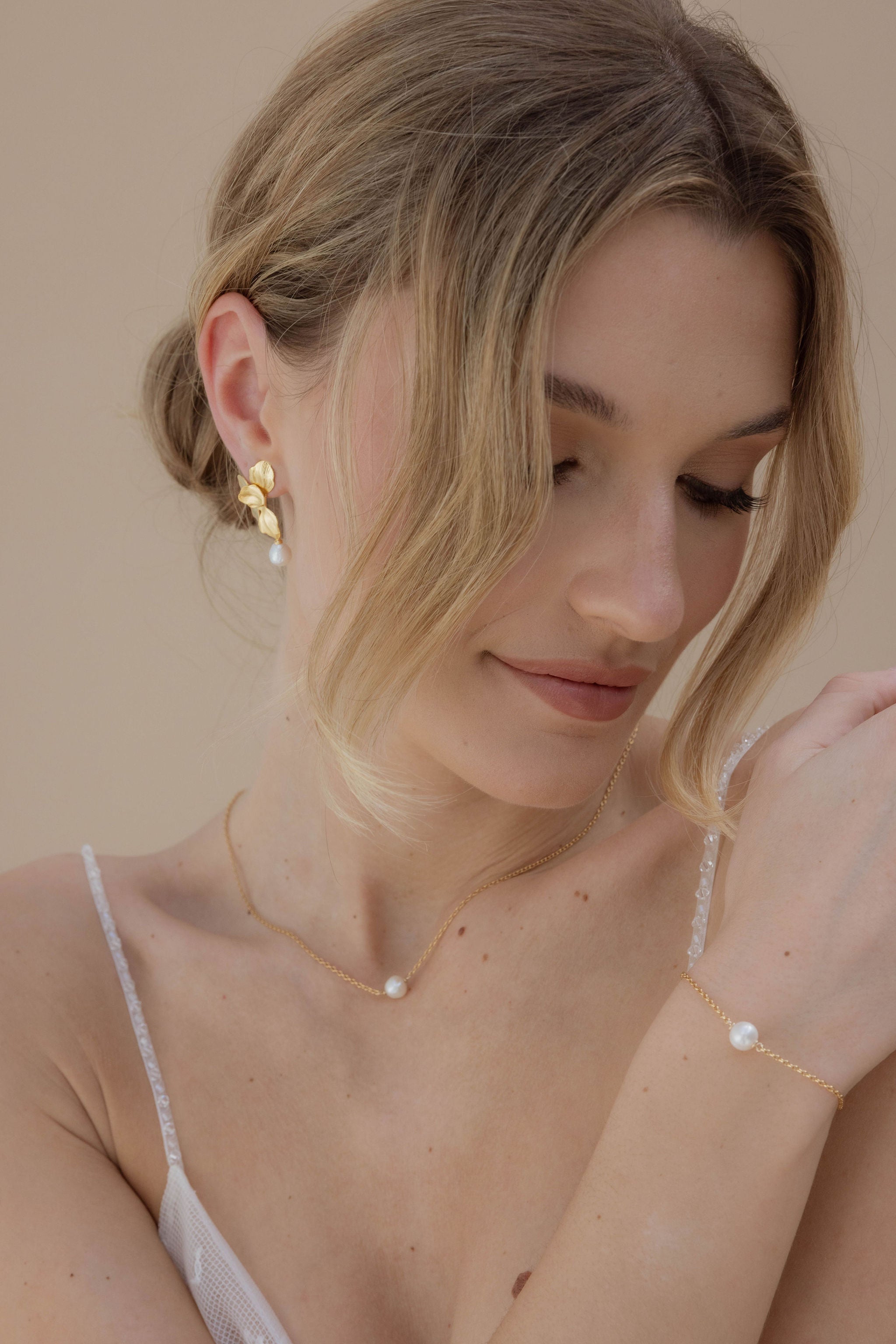 Mariée qui regarde vers le bas avec des bijoux dores ornes de perles, elle porte un bracelet, un collier et des boucles d'oreilles assorties.