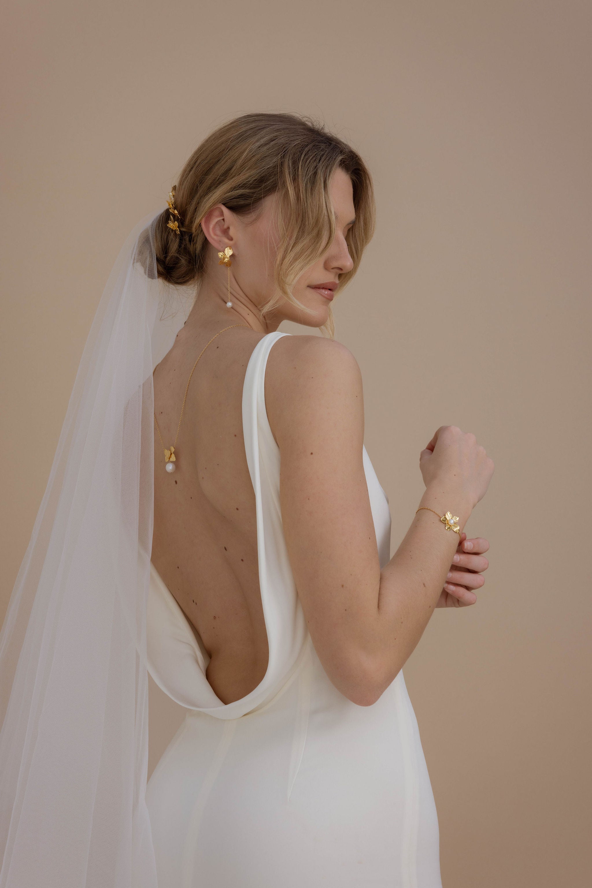 Mariée coiffée d’un chignon bas orné de pics fleuris, portant un collier de dos raffiné et des boucles pendantes. Dans une jolie robe avec dos nu et un voile.