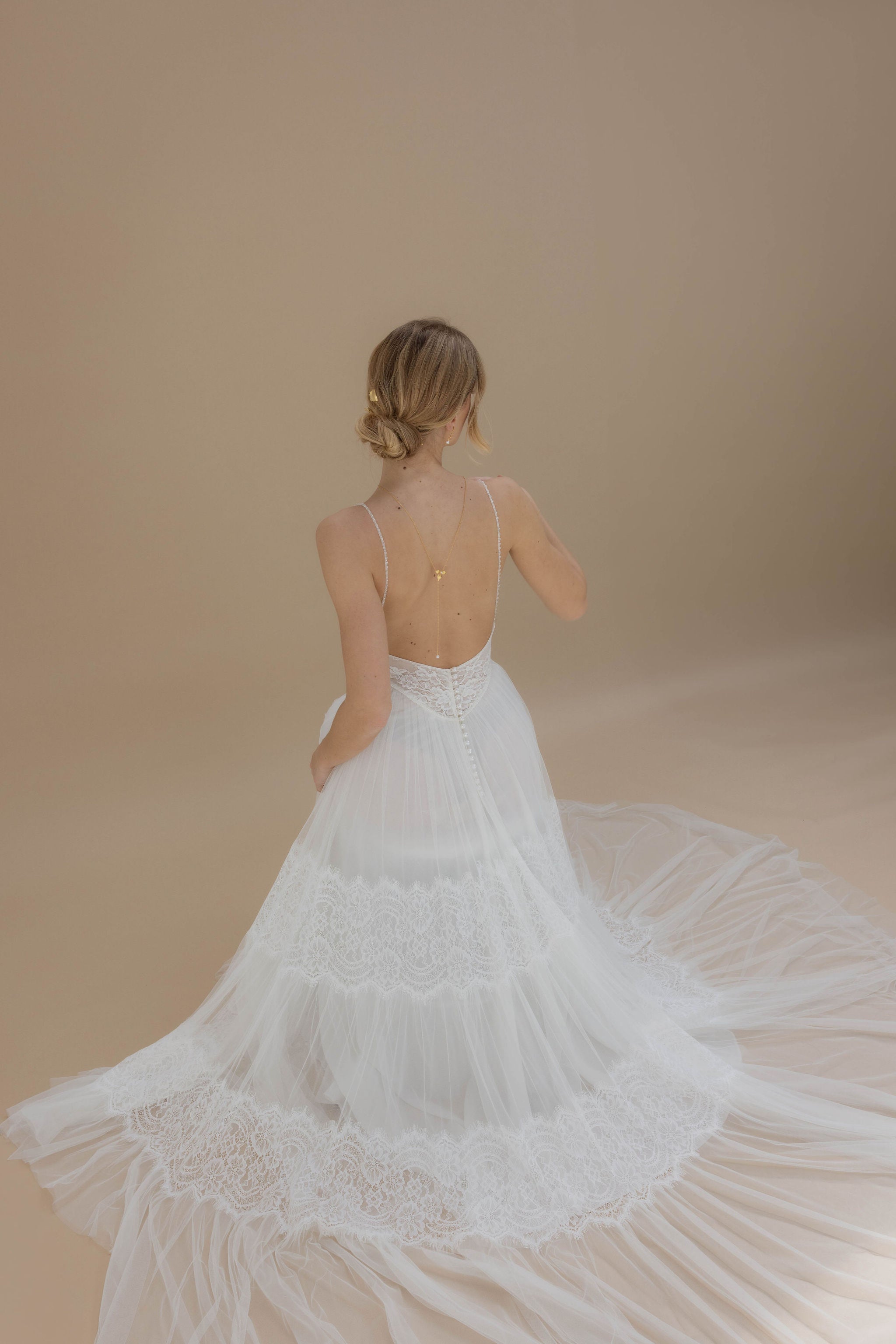 Vue arrière d’une mariée assise avec robe ample, chignon bas soigné, et collier de dos délicat, parfait pour un mariage romantique.