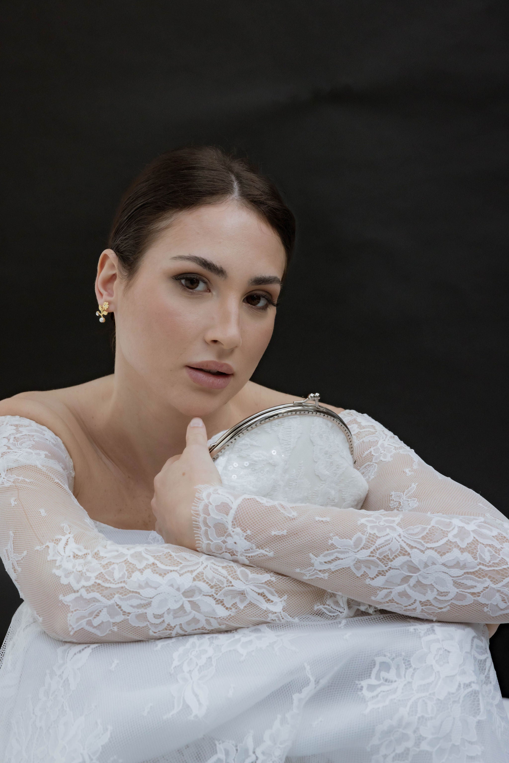 Mariée en robe blanche à dentelle, tenant un sac blanc avec un arc argenté, regard droit devant, portant de jolies boucles d’oreilles dorées et un chignon bien attaché.