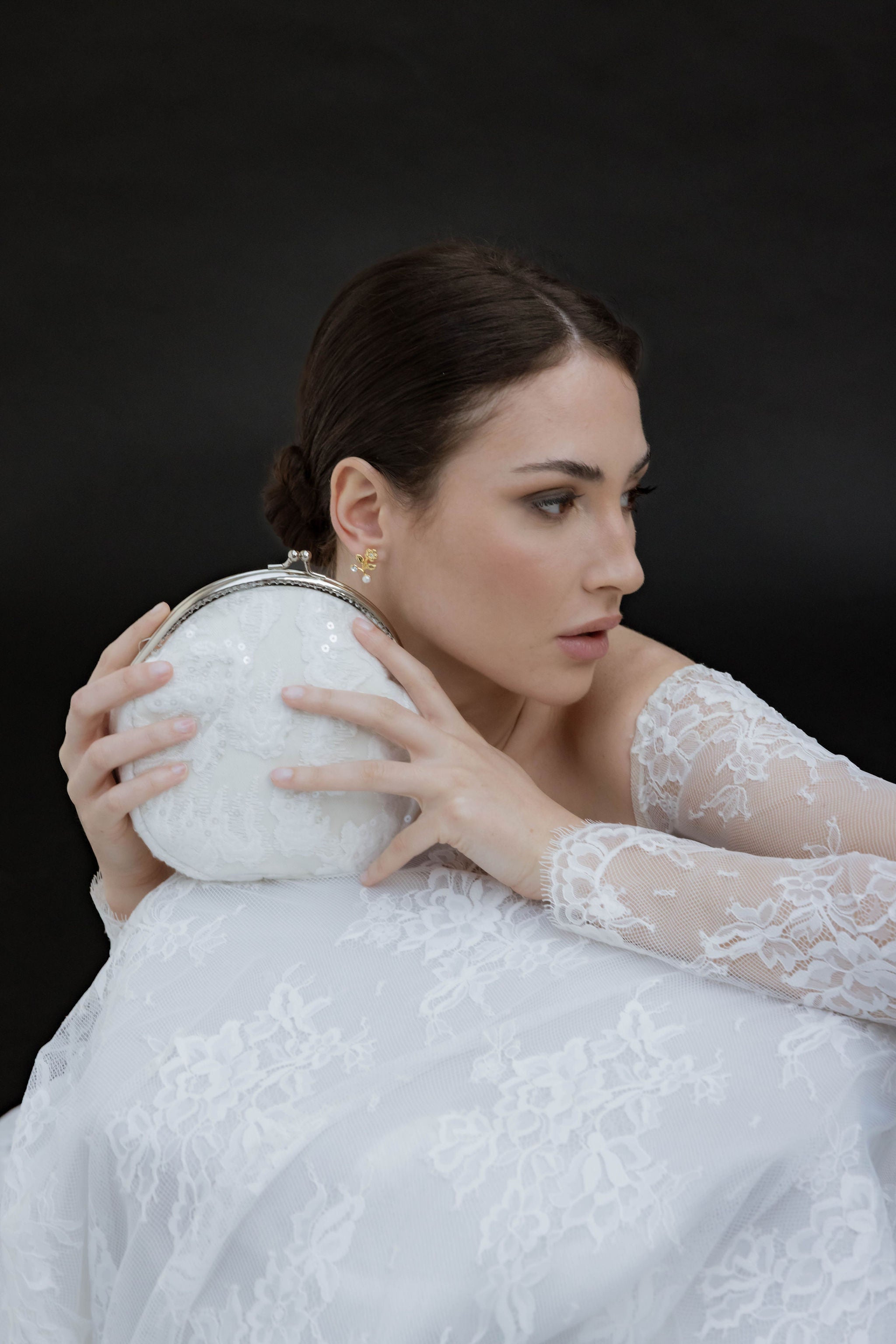 Mariée en robe blanche à dentelle, regardant sur le côté, tenant son sac blanc avec un arc argenté à côté de sa tête, portant de jolies boucles d’oreilles dorées et un chignon bien attaché.