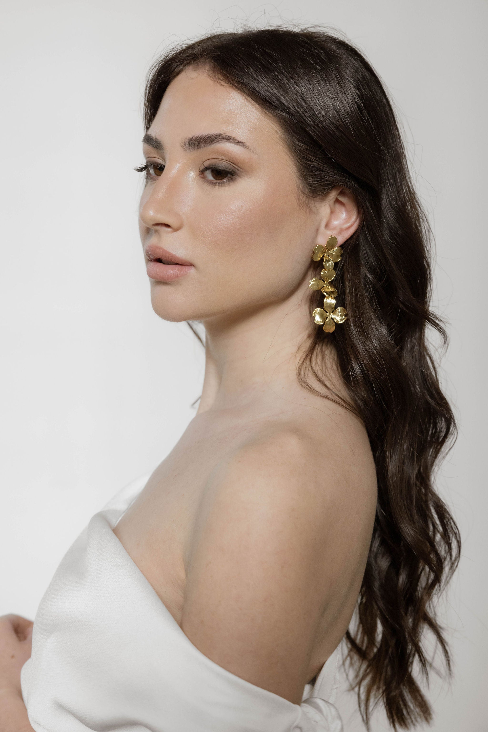 Mariée en robe blanche bustier, les cheveux longs et ondulés, portant de longues boucles d’oreilles dorées florales qui ajoutent une touche romantique.