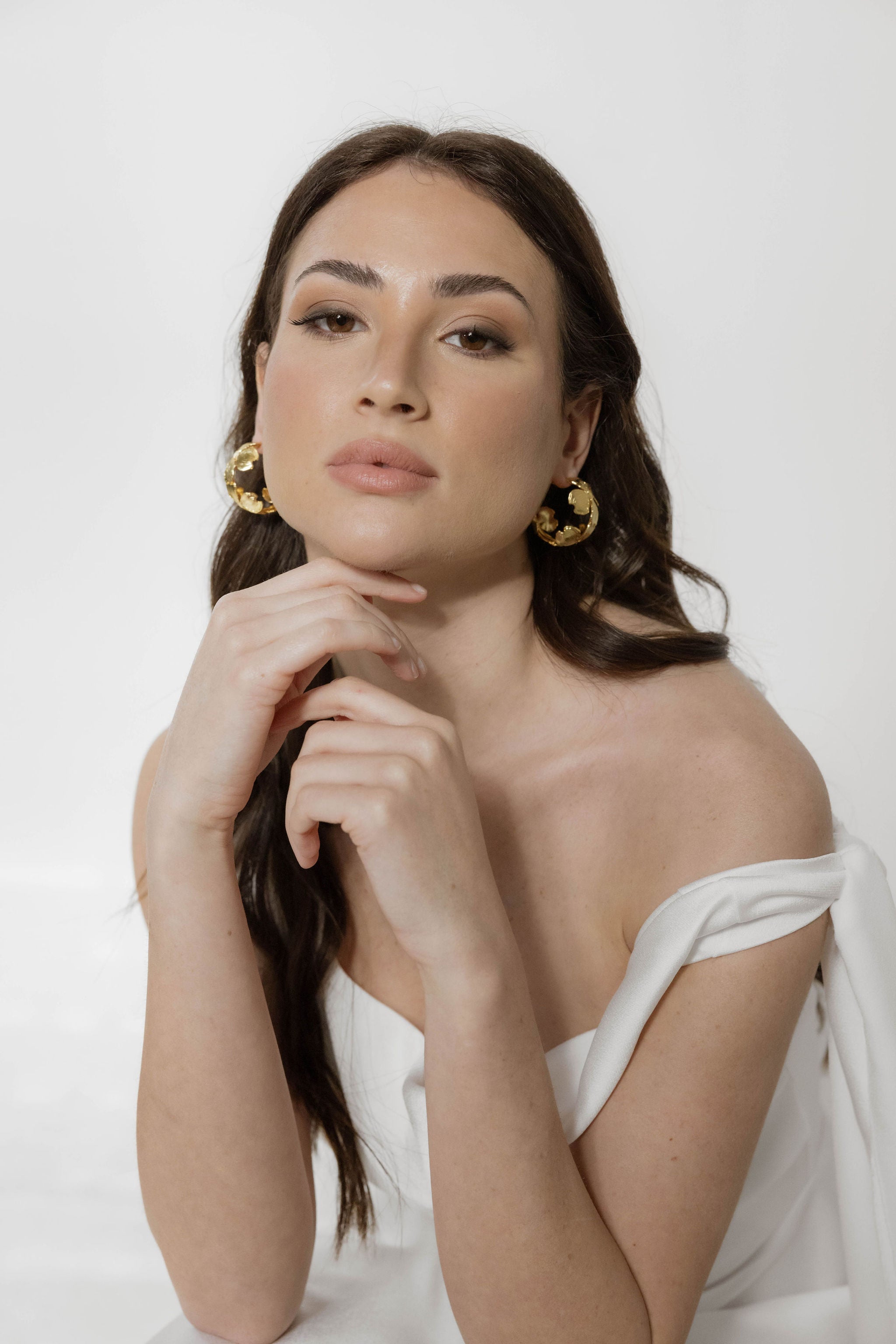 Mariée en robe bustier, cheveux ondulés, reposant sa tête sur sa main, portant de grandes créoles fleuries qui ajoutent une touche de délicatesse à son look.