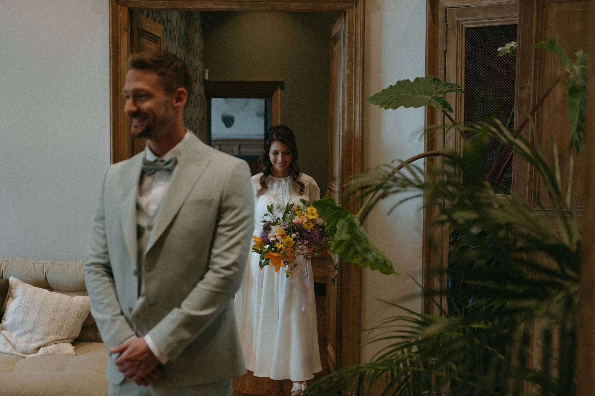 Photo mariage élégant, mariée portant bouquet de fleurs colorées derrière mari en costume souriant, robe de mariée deux pièces élégante, couple dans pièce de château aux portes en bois, inspiration mariage château