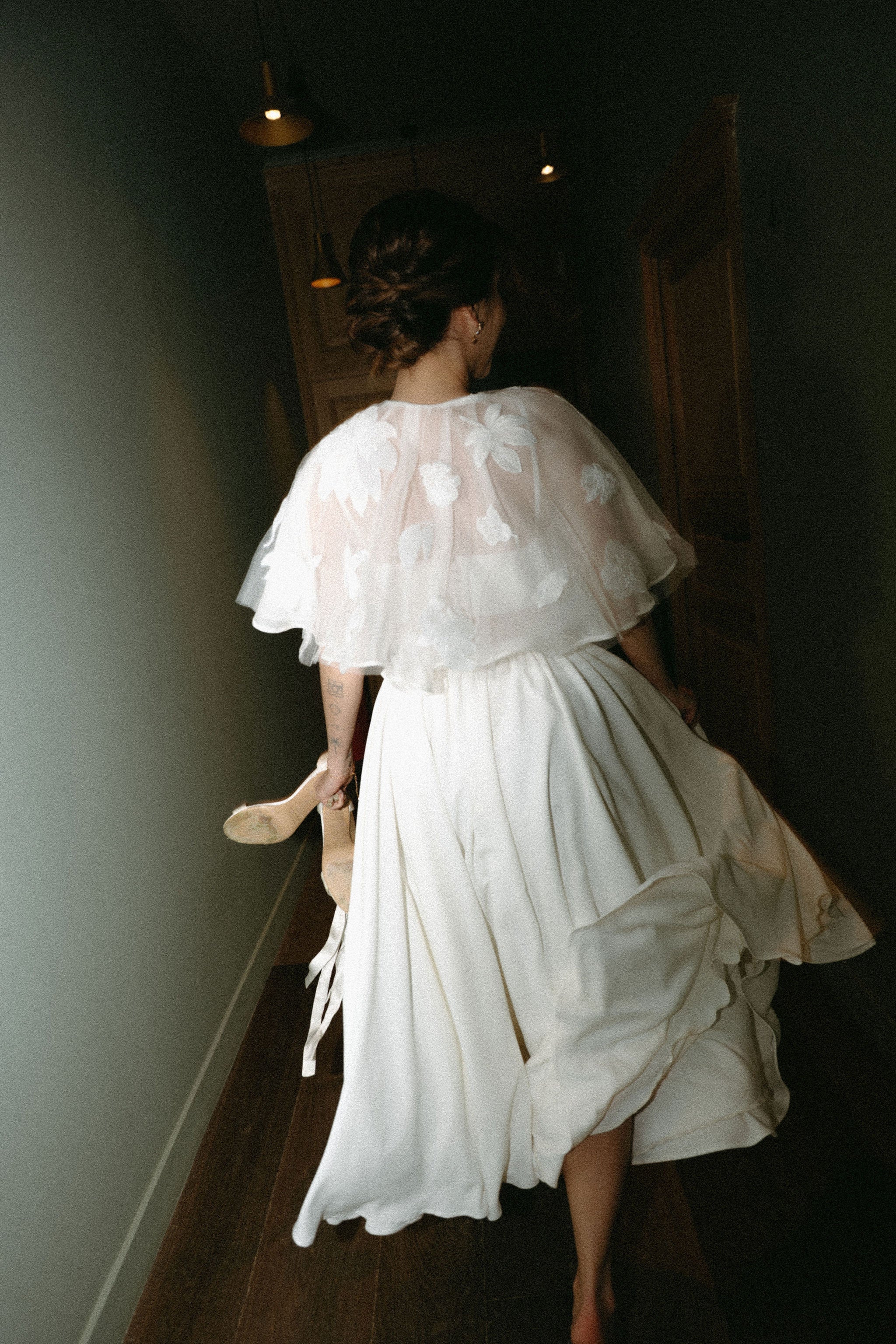 Mariée de dos courant dans couloir, tenant talons à la main, robe de mariée deux pièces élégante, châle en dentelle, tenue de mariage raffinée, photo mariage originale