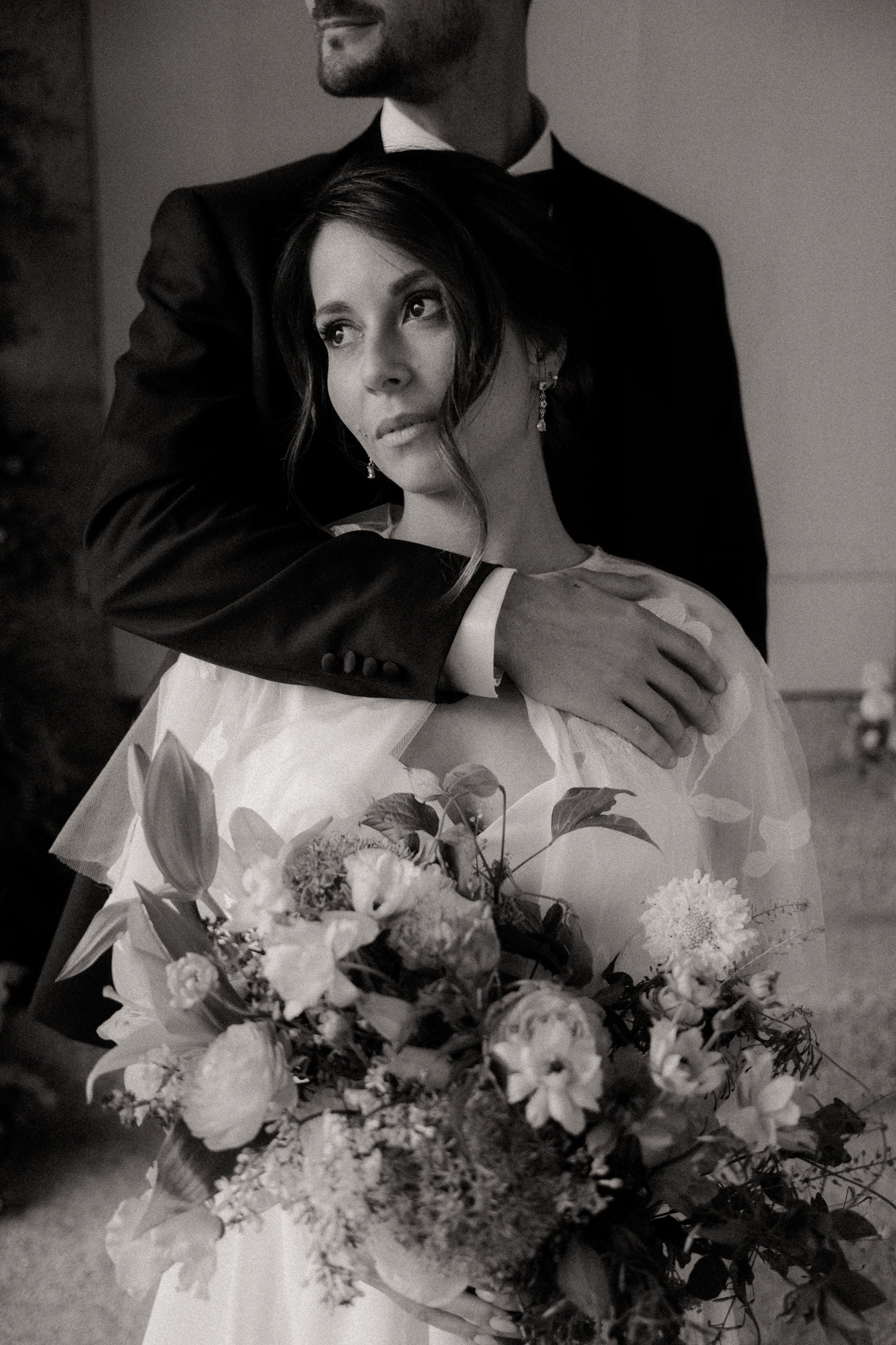 Photo en noir et blanc d'un couple marié, homme en costume qui enlace sa femme en robe de mariée, mariée élégante qui tient un bouquet de fleurs et qui porte les boucles d'oreilles vitoria Atelier Lilac