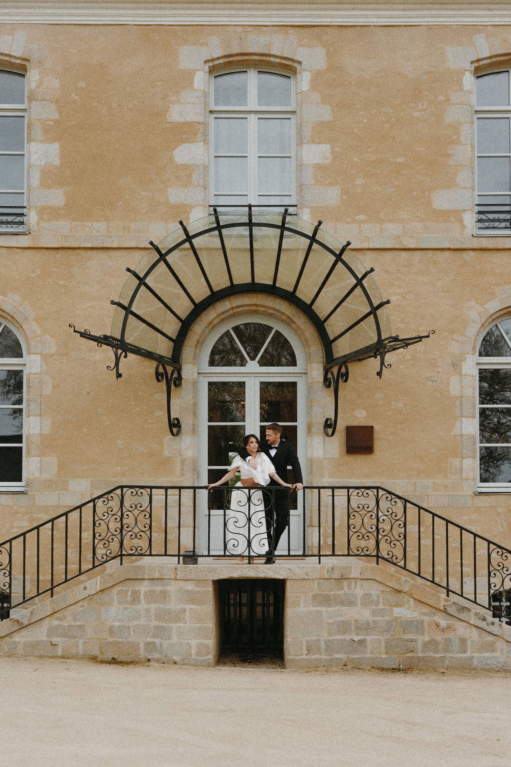 Couple marié élégant dans les bras l’un de l’autre devant porte de château, au-dessus de deux escaliers, mariée en robe deux pièces, marié en costume