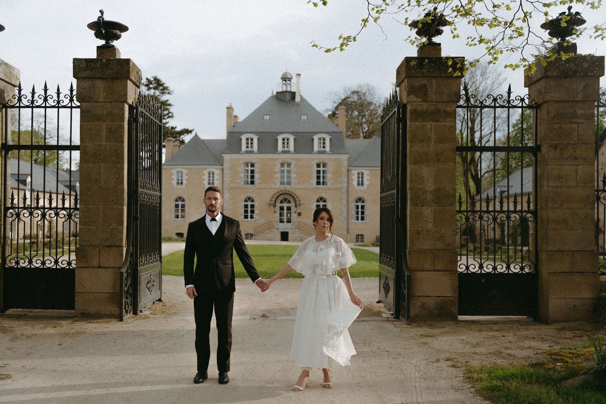 Photo mariage château. Couple marié élégant se tenant la main devant château, mariée en robe deux pièces, marié en costume, mariage romantique au château